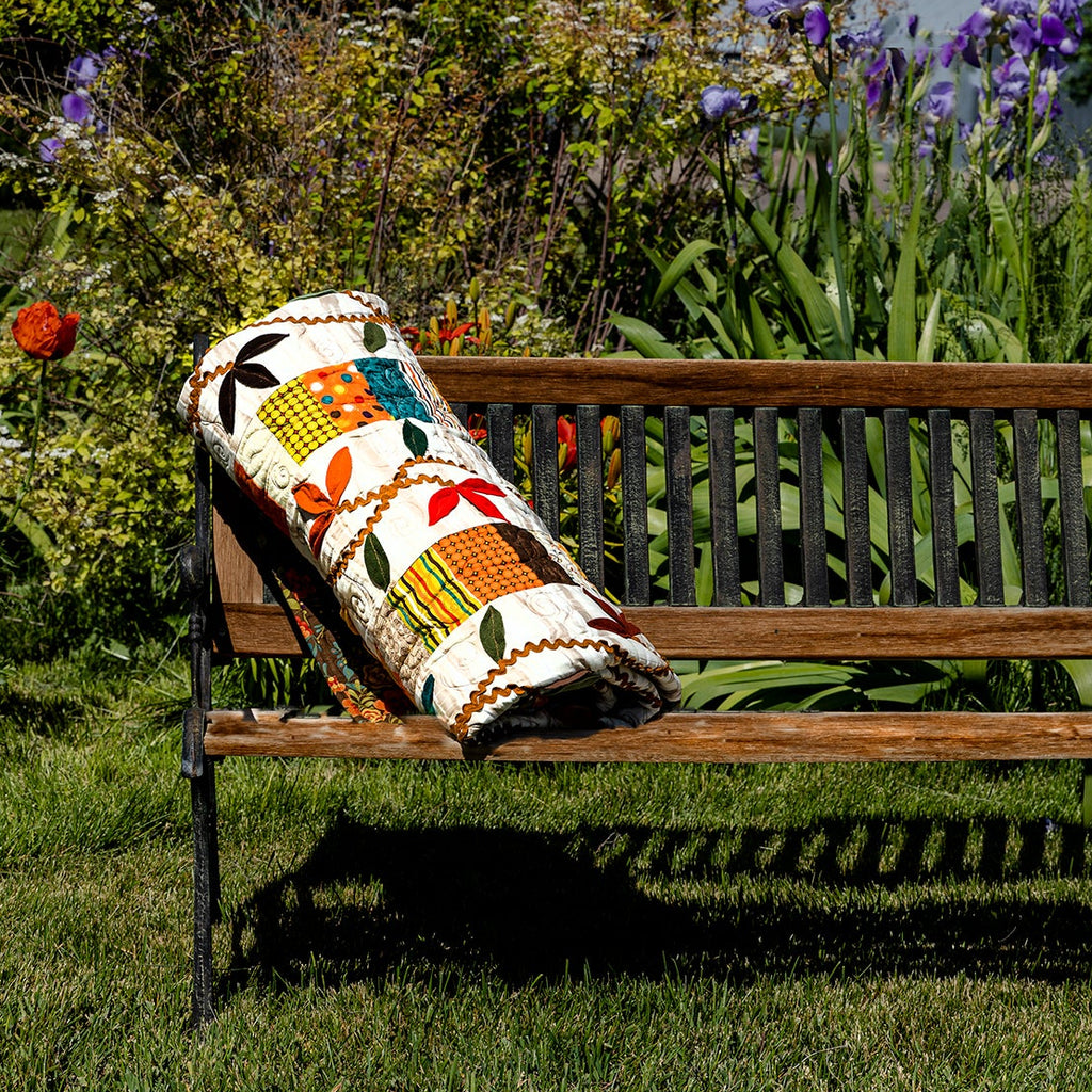 A colorful woven throw pillow with a geometric pattern rests on a wooden park bench in a lush green garden with purple flowers.