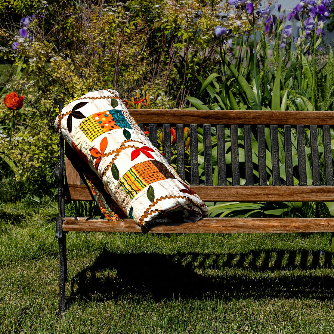 A colorful woven throw pillow with a geometric pattern rests on a wooden park bench in a lush green garden with purple flowers.