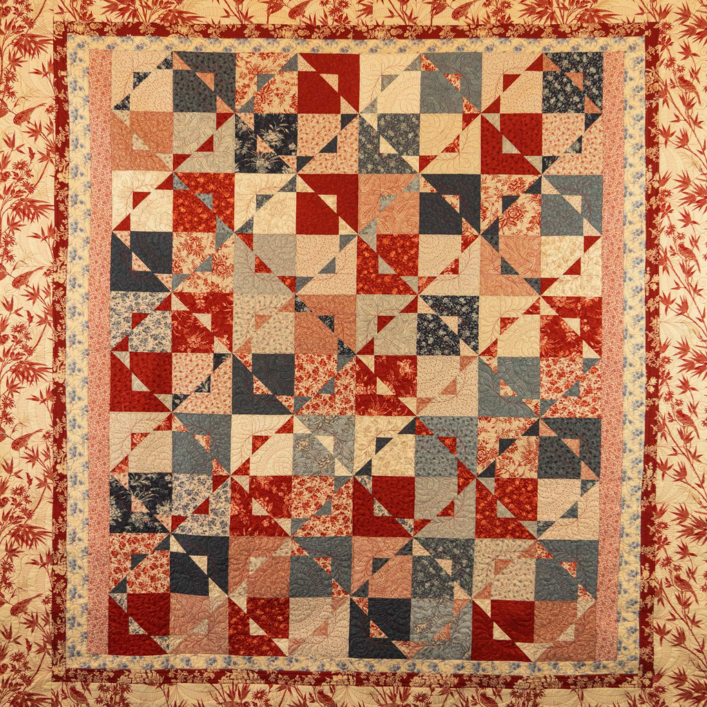 A vintage-style patchwork quilt with a geometric starburst pattern in red, beige, navy, and gray, surrounded by a red floral border.