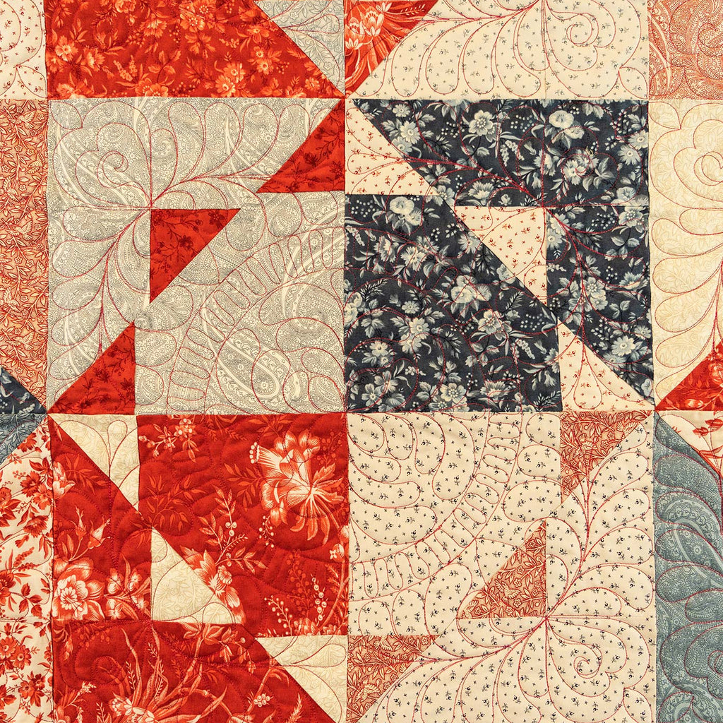 A quilted fabric panel featuring a geometric patchwork design with red, cream, navy floral, and muted blue patterned sections, all decorated with swirling quilting stitches.