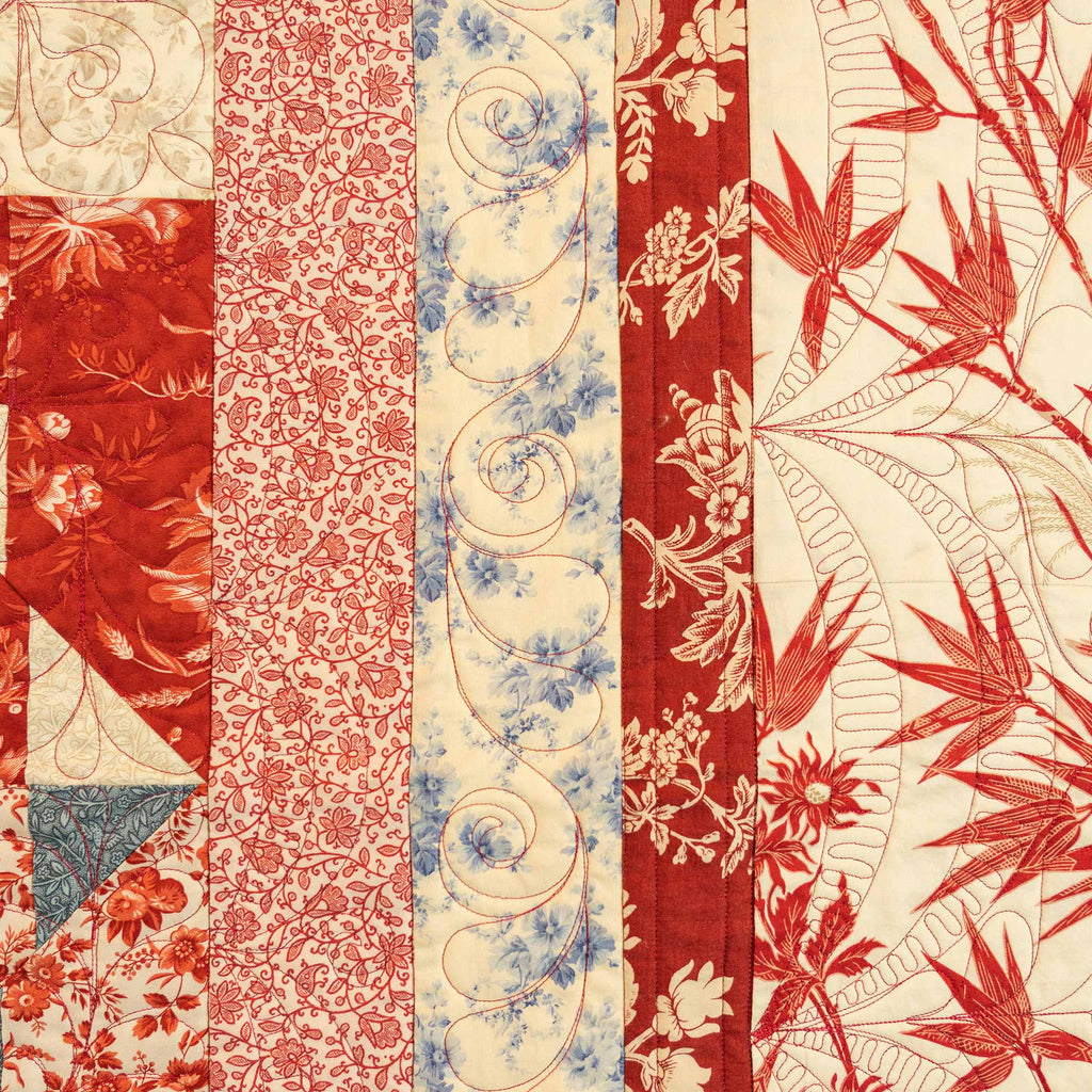 A close-up of a vintage quilt or textile panel featuring vertical stripes of red, coral, cream, and blue floral and scroll patterns.
