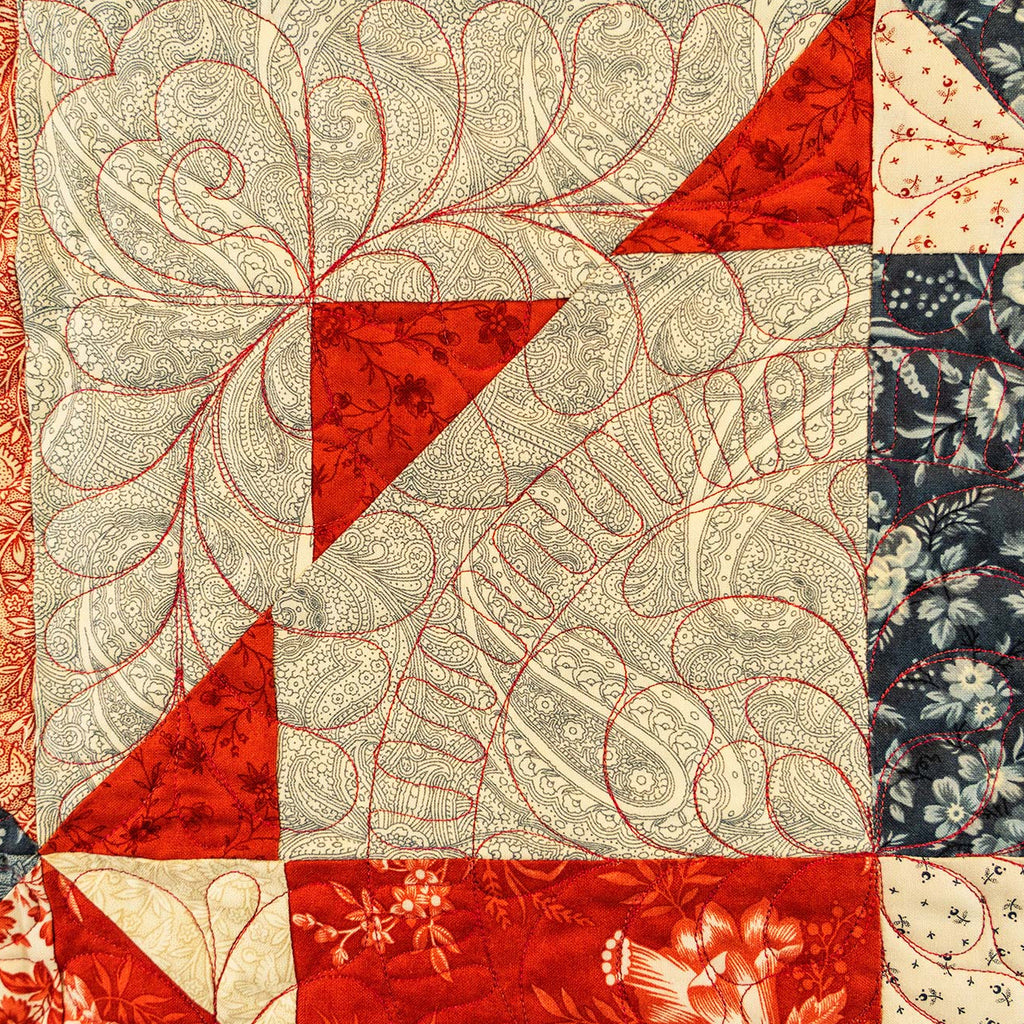 A close-up of a quilted textile with a patchwork design featuring red, cream, and navy patterned fabric sections, including paisley, floral, and leaf motifs.