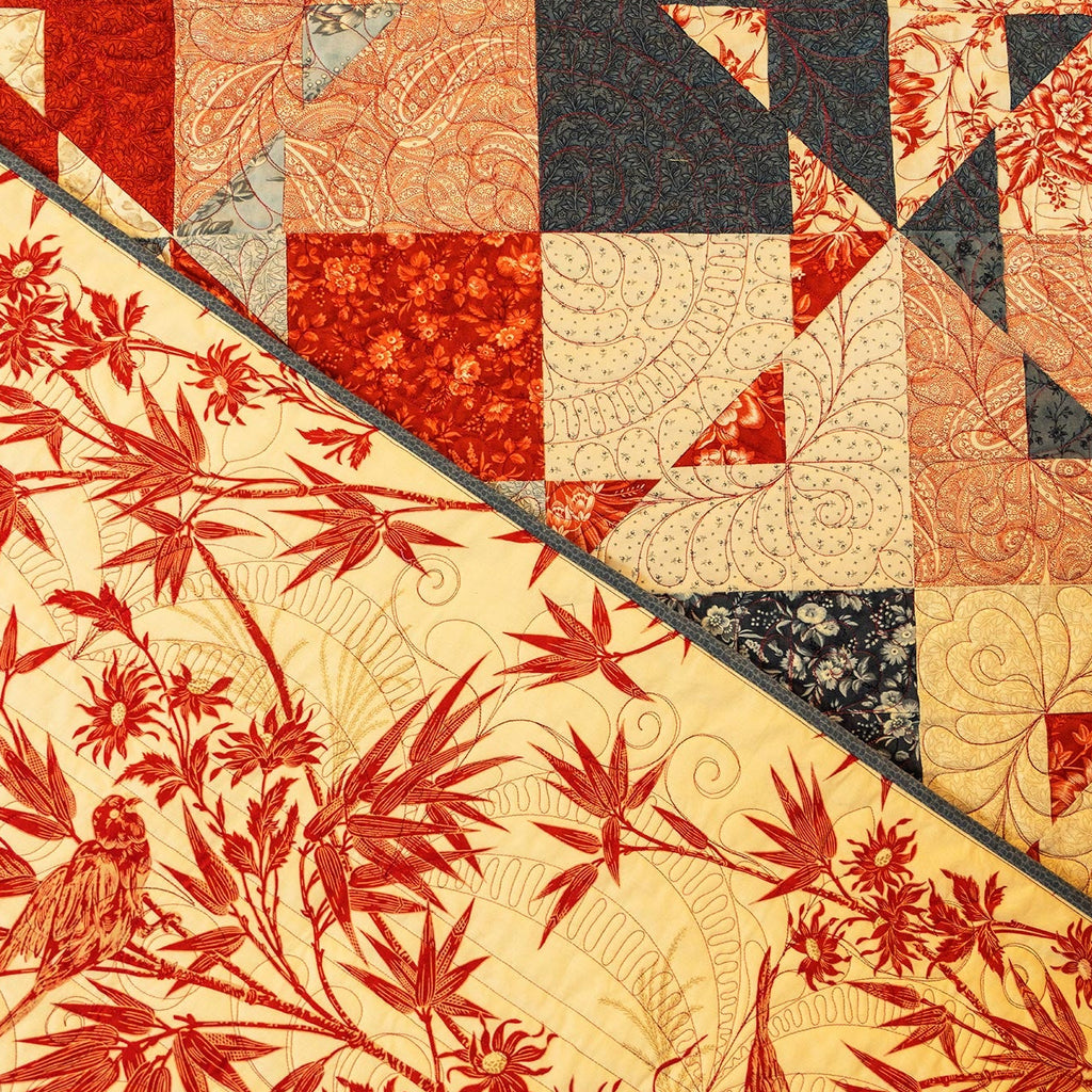 A close-up of a quilted fabric featuring a diagonal split design: one side shows a red botanical and bird print on a cream background, and the other side shows a patchwork of red, beige, and dark gray quilted fabric blocks.