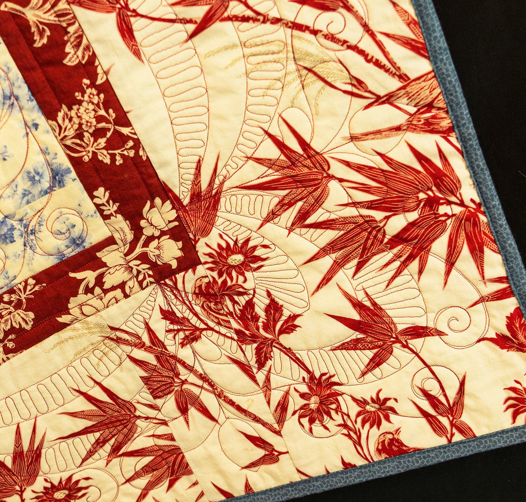 A close-up of a quilted textile featuring a red and beige botanical pattern with visible quilting stitches and a dark blue border, shown against a black background.