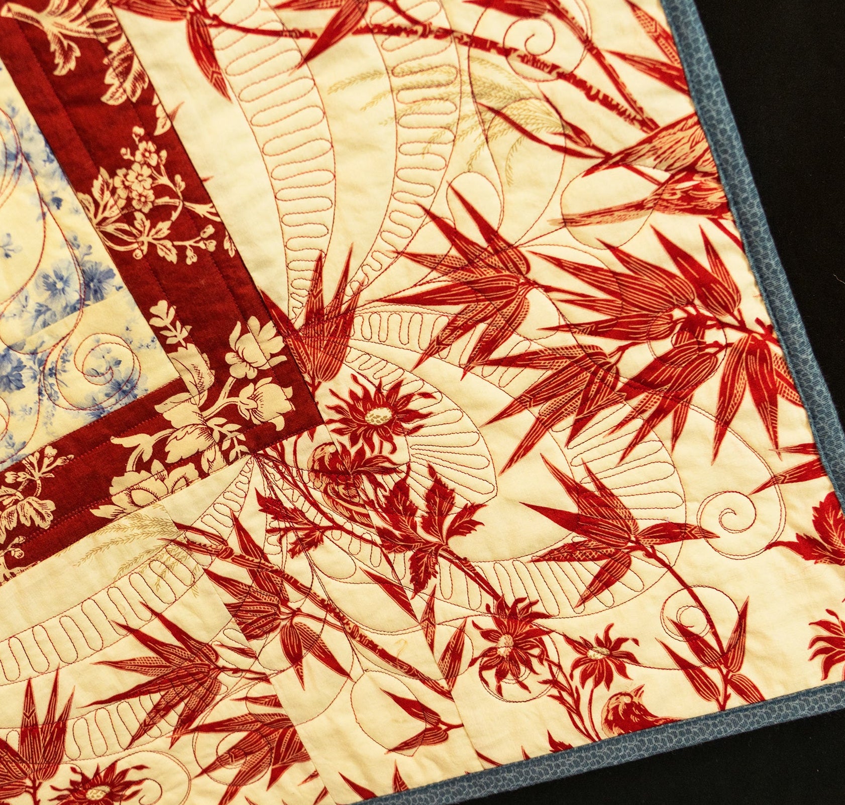 A close-up of a quilted textile featuring a red and beige botanical pattern with visible quilting stitches and a dark blue border, shown against a black background.