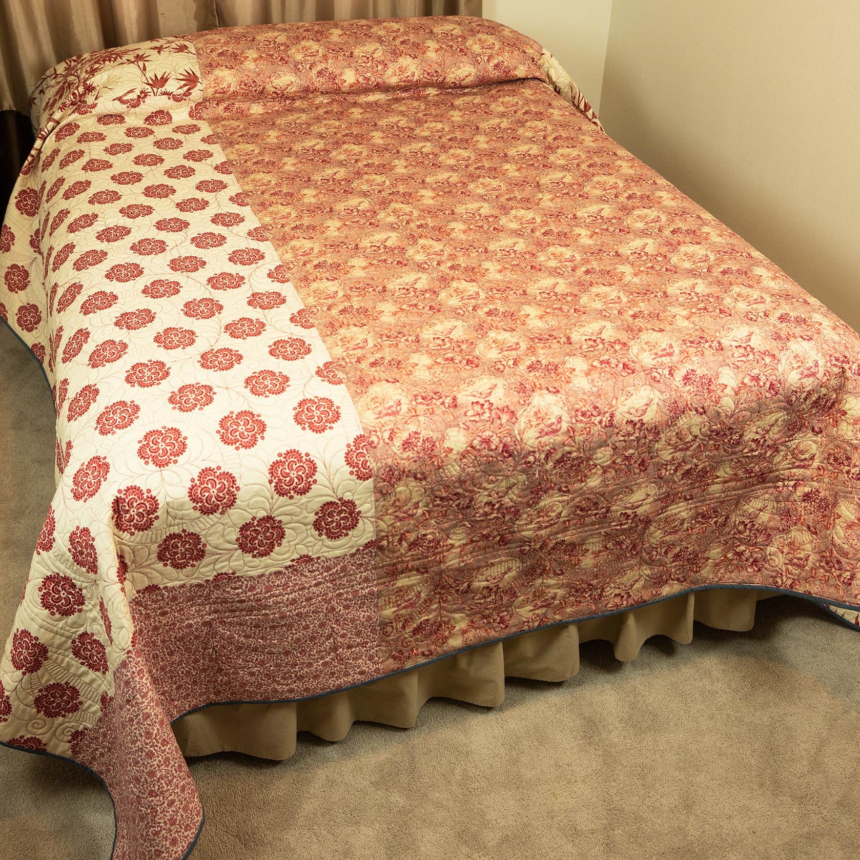 The reverse of a patchwork quilted bedspread with a floral pattern in muted red and beige, draped over a bed with a beige ruffled bed skirt in a bedroom setting.