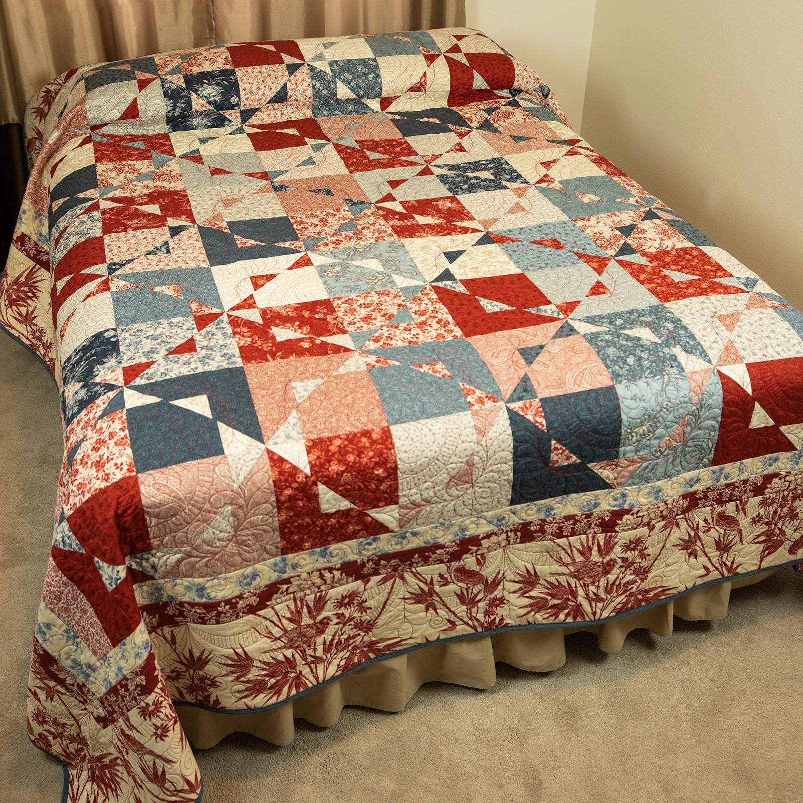 A patchwork quilted bedspread with a matching ruffled bed skirt, featuring geometric and floral blocks in red, navy blue, light blue, and cream, styled on a bed in a bedroom.