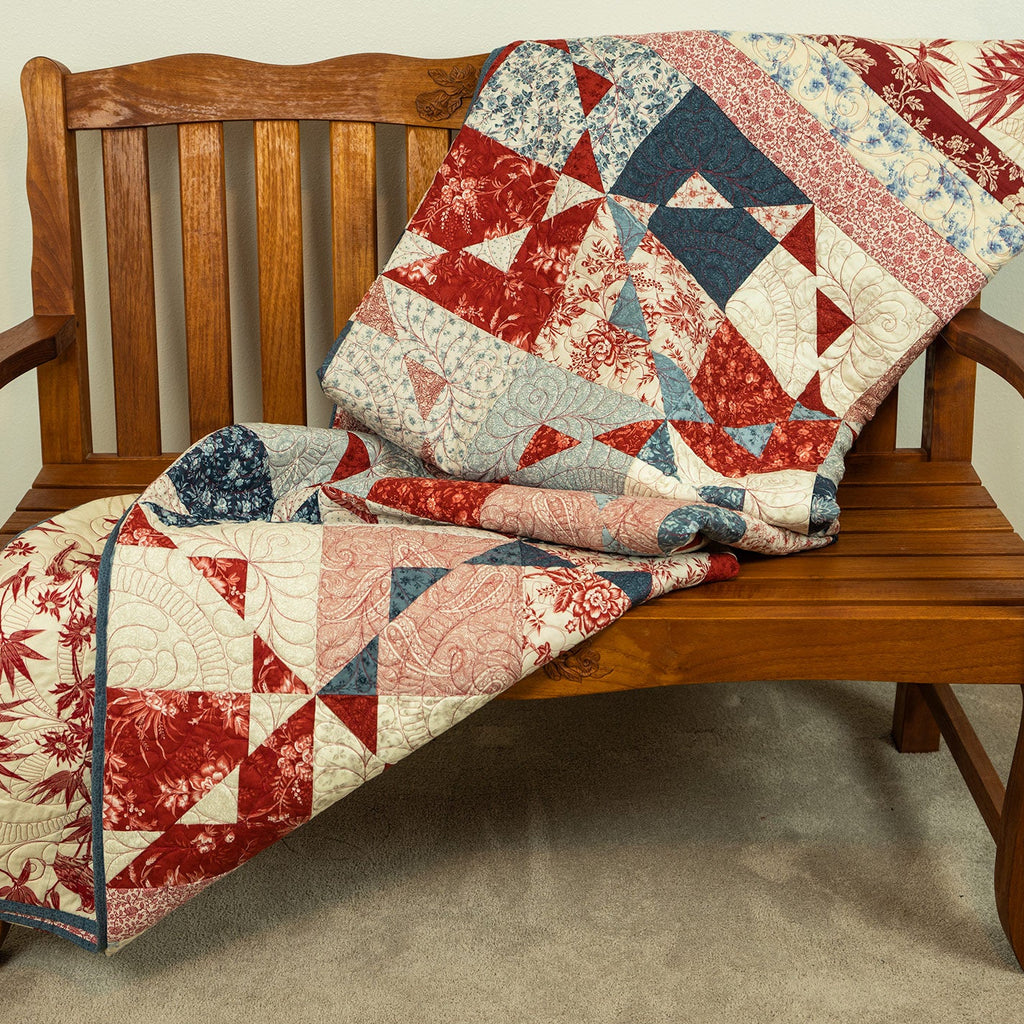 A patchwork quilt with red, blue, and cream floral and geometric patterns draped over a wooden bench in an indoor setting.