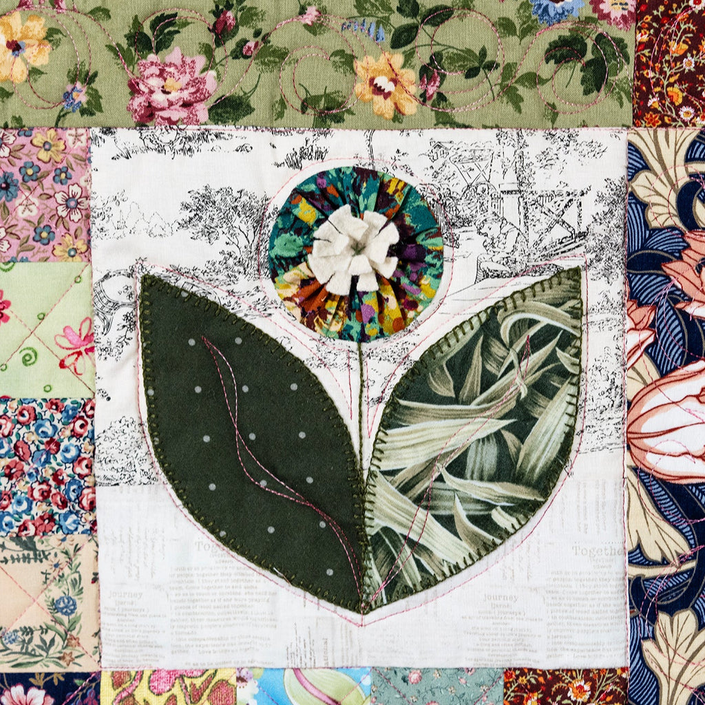 A close-up of a patchwork quilt featuring a central floral applique with two large green leaves and a multicolored circular flower, surrounded by various patterned fabric borders.