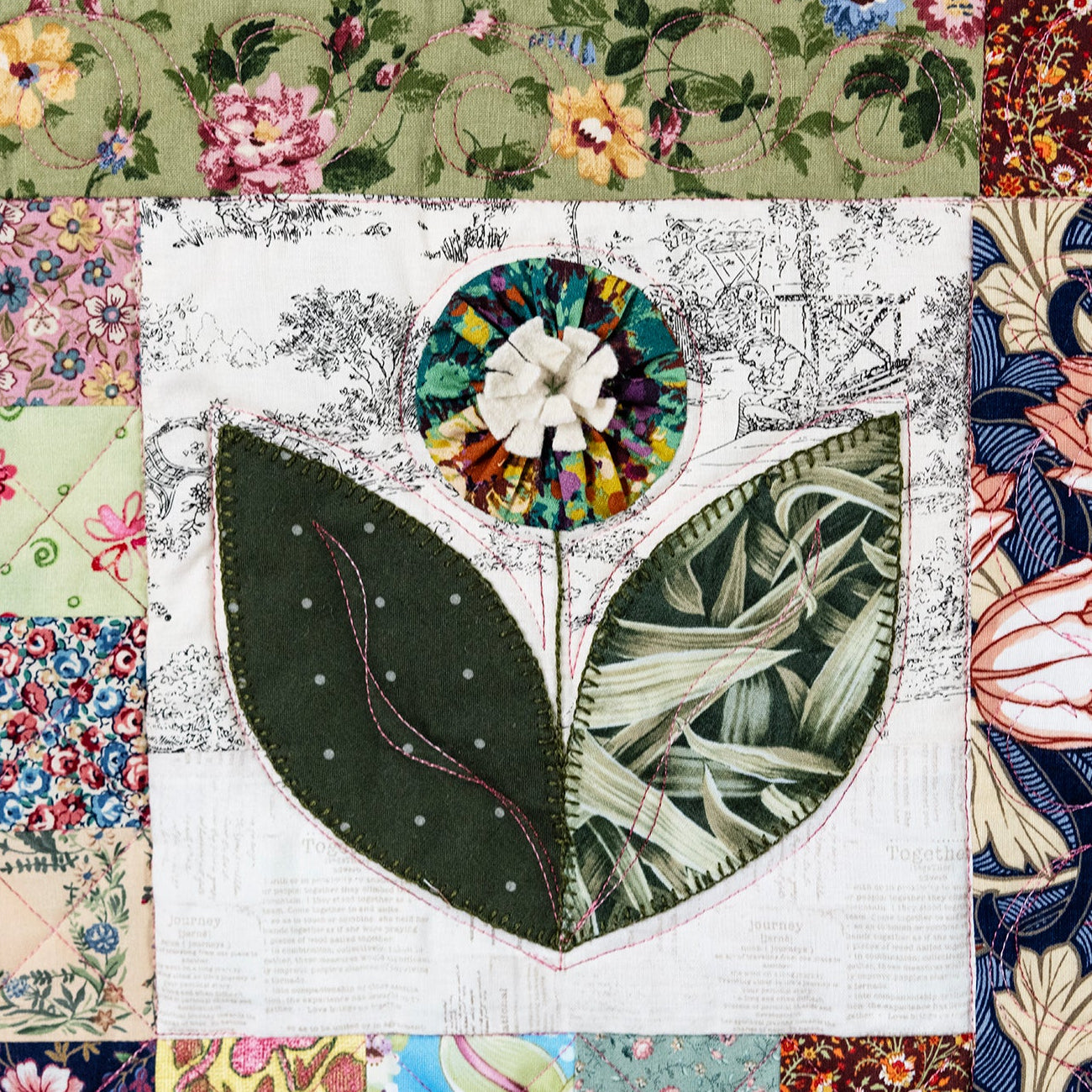A close-up of a patchwork quilt featuring a central floral applique with two large green leaves and a multicolored circular flower, surrounded by various patterned fabric borders.