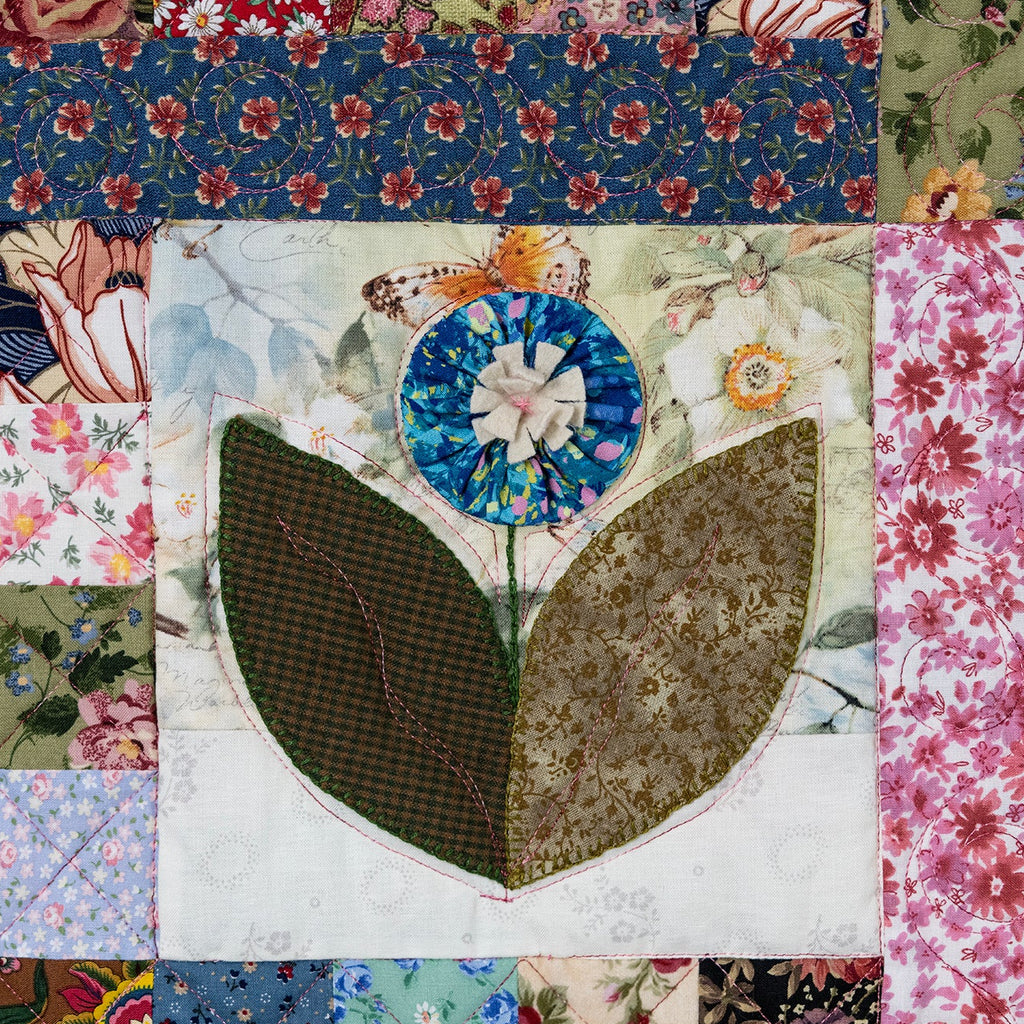 A square patchwork quilt featuring a central floral motif with two large olive-green leaves and a blue flower, surrounded by various vintage floral and botanical fabric panels.