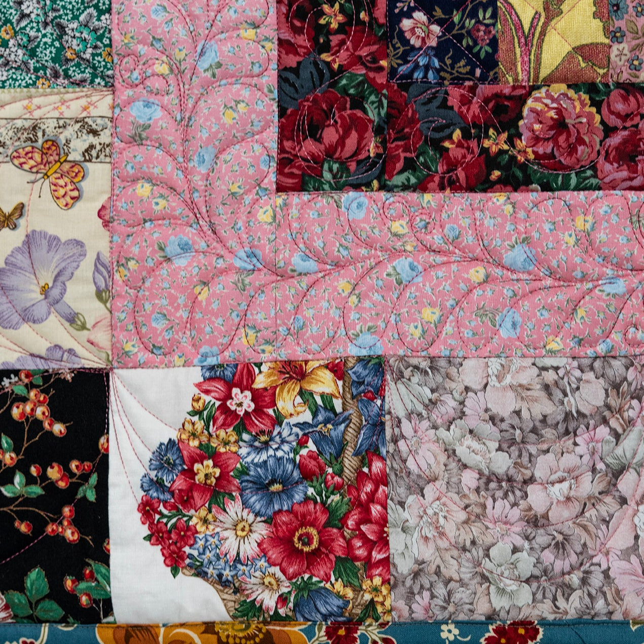 A square patchwork quilt featuring multiple floral and botanical fabric panels in pink, red, green, black, white, and purple, stitched together with a decorative border.