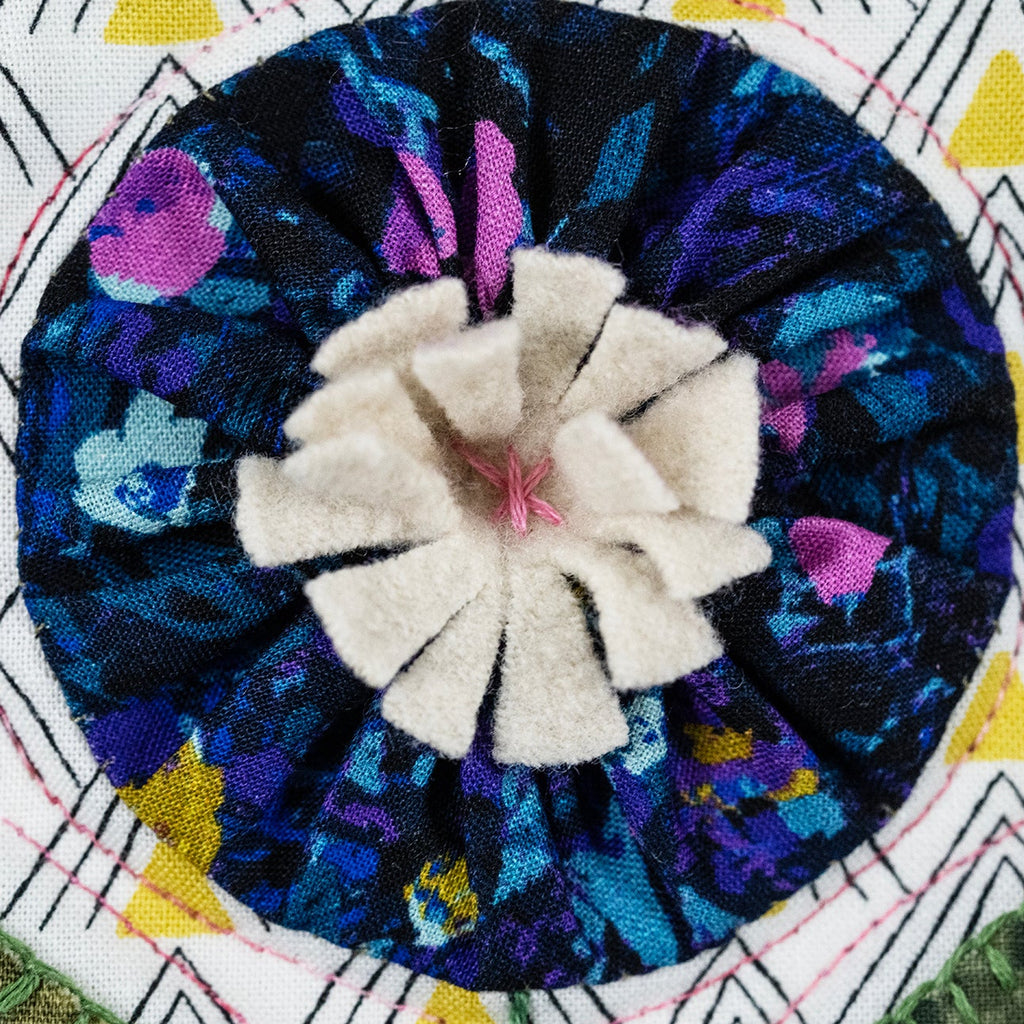 A close-up of a circular fabric applique or brooch featuring a white felt flower center with a dark blue and purple patterned fabric rosette, set against a white quilted background with geometric yellow and black patterns.