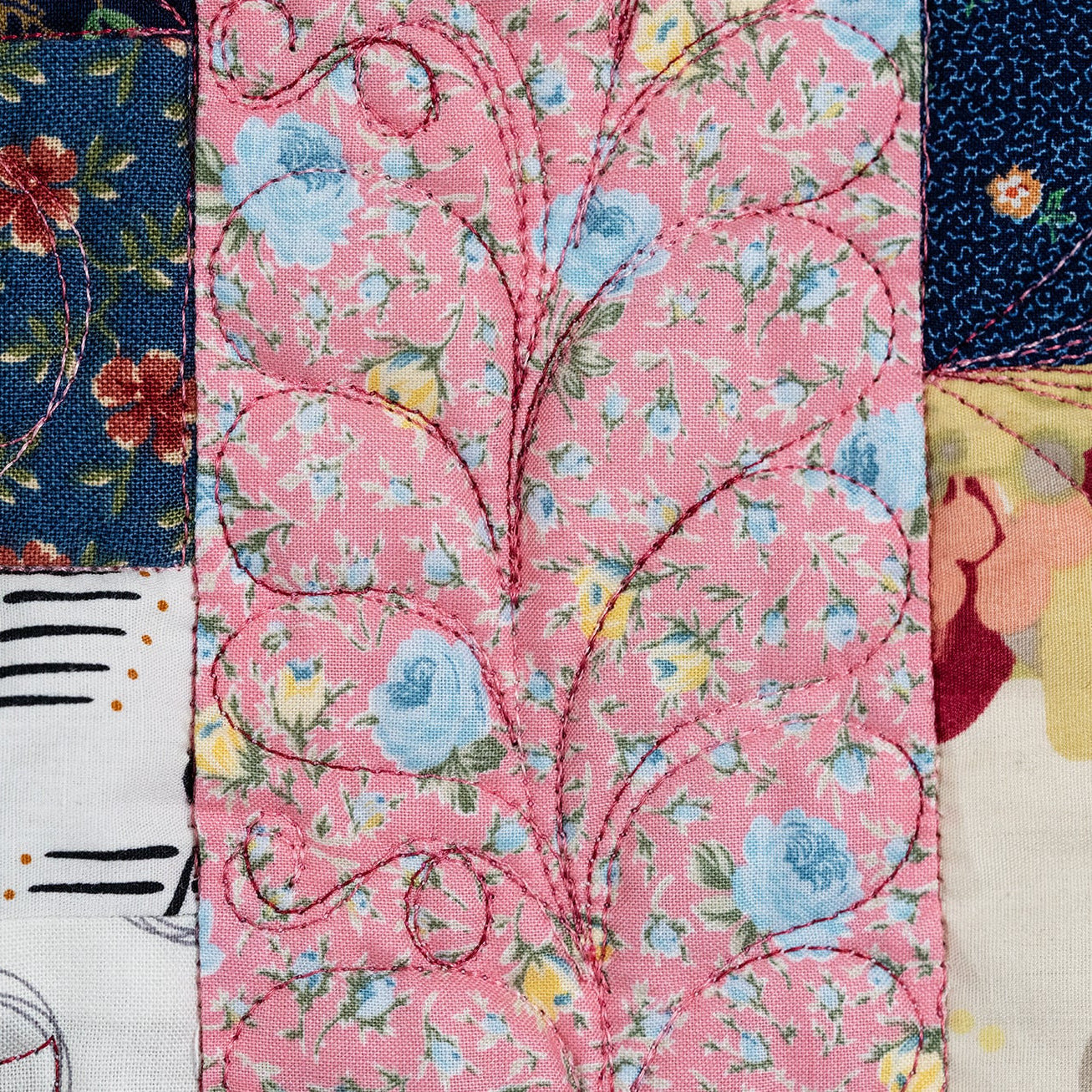 A close-up of a quilted textile featuring three vertical panels with different floral and abstract patterns: a navy blue panel with red flowers and green leaves, a pink panel with blue and yellow floral motifs and swirling stitched lines, and a cream panel with red, yellow, and gray abstract shapes.