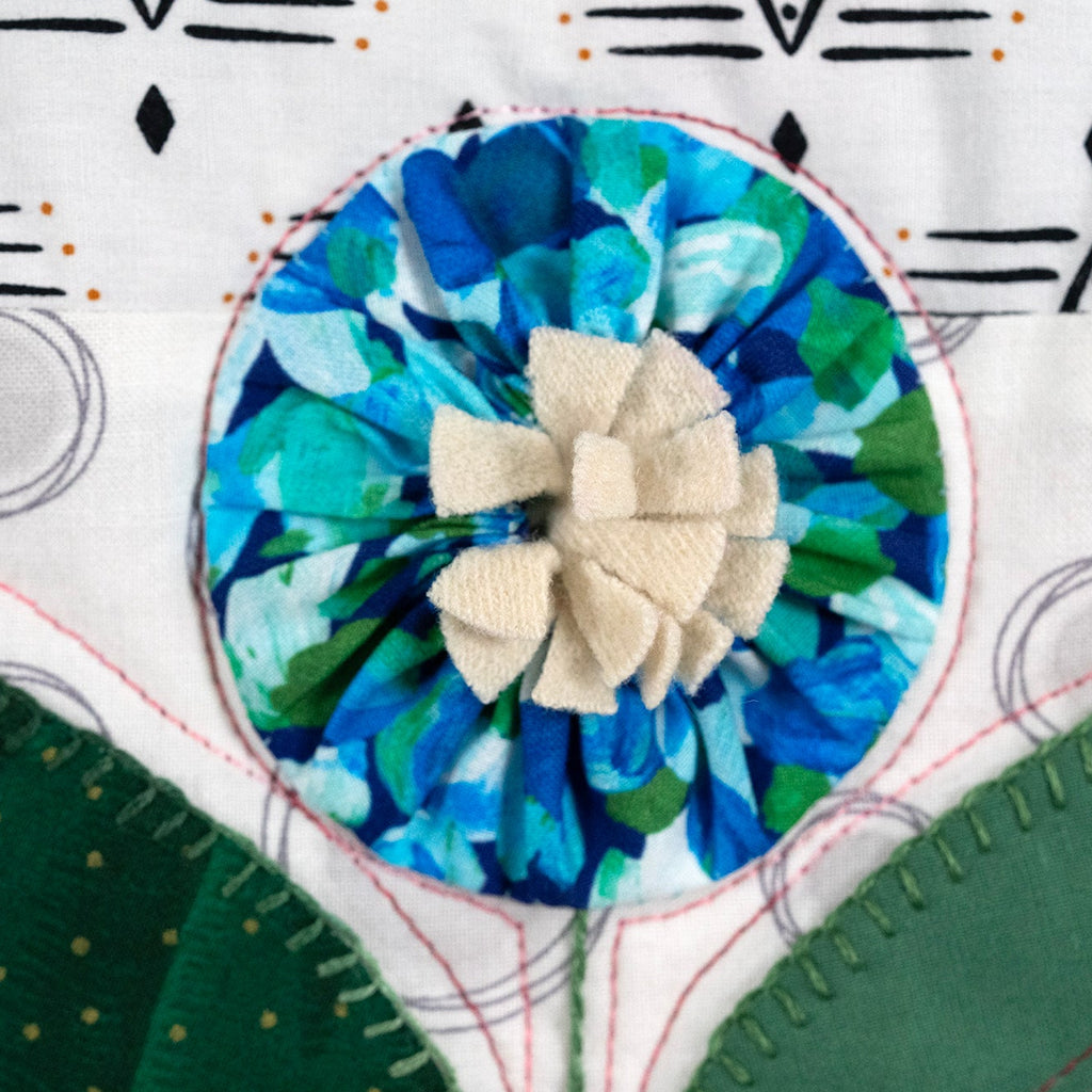 Close-up of a handmade quilted fabric design featuring a central circular floral applique with blue and green patterned petals and a beige fabric flower in the center, surrounded by white and green quilted sections with decorative stitching.