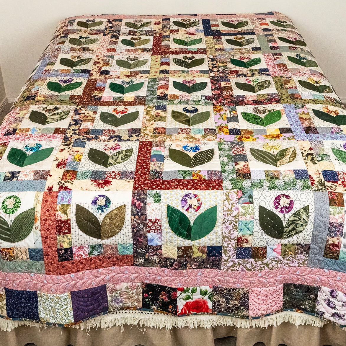 A multicolor patchwork quilt with leaf and floral motifs, draped over a bed with a beige ruffled bed skirt in a bedroom setting.