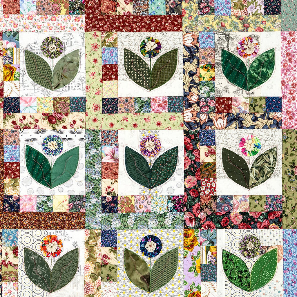 A square quilted textile featuring a grid of floral and leaf appliqué blocks in many colors, with cream, green, pink, blue, and brown patterned fabrics visible across the surface.