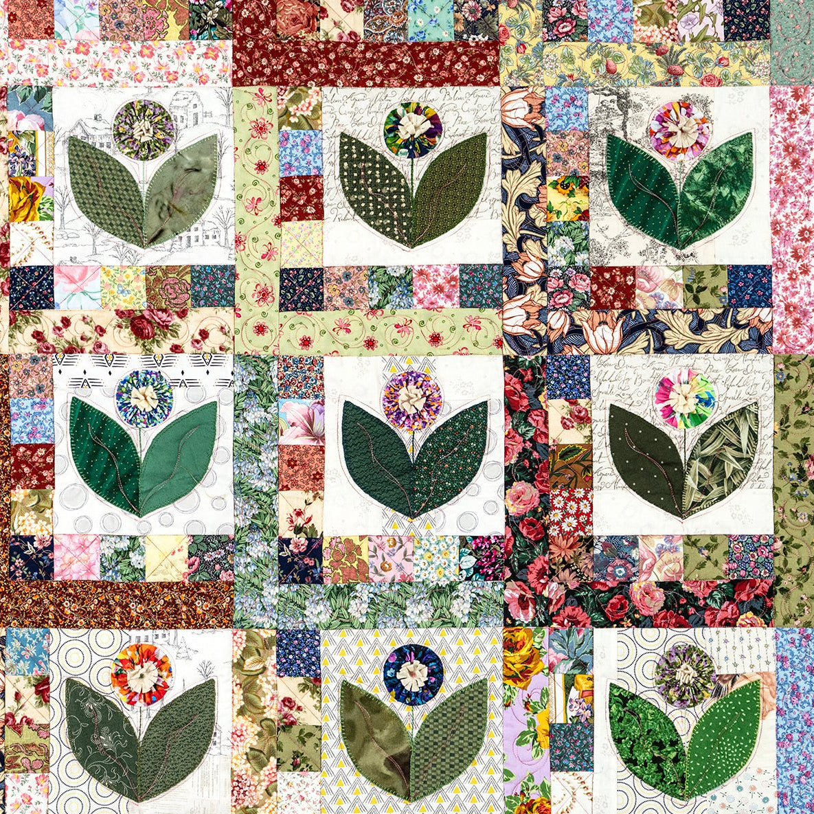 A square quilted textile featuring a grid of floral and leaf appliqué blocks in many colors, with cream, green, pink, blue, and brown patterned fabrics visible across the surface.