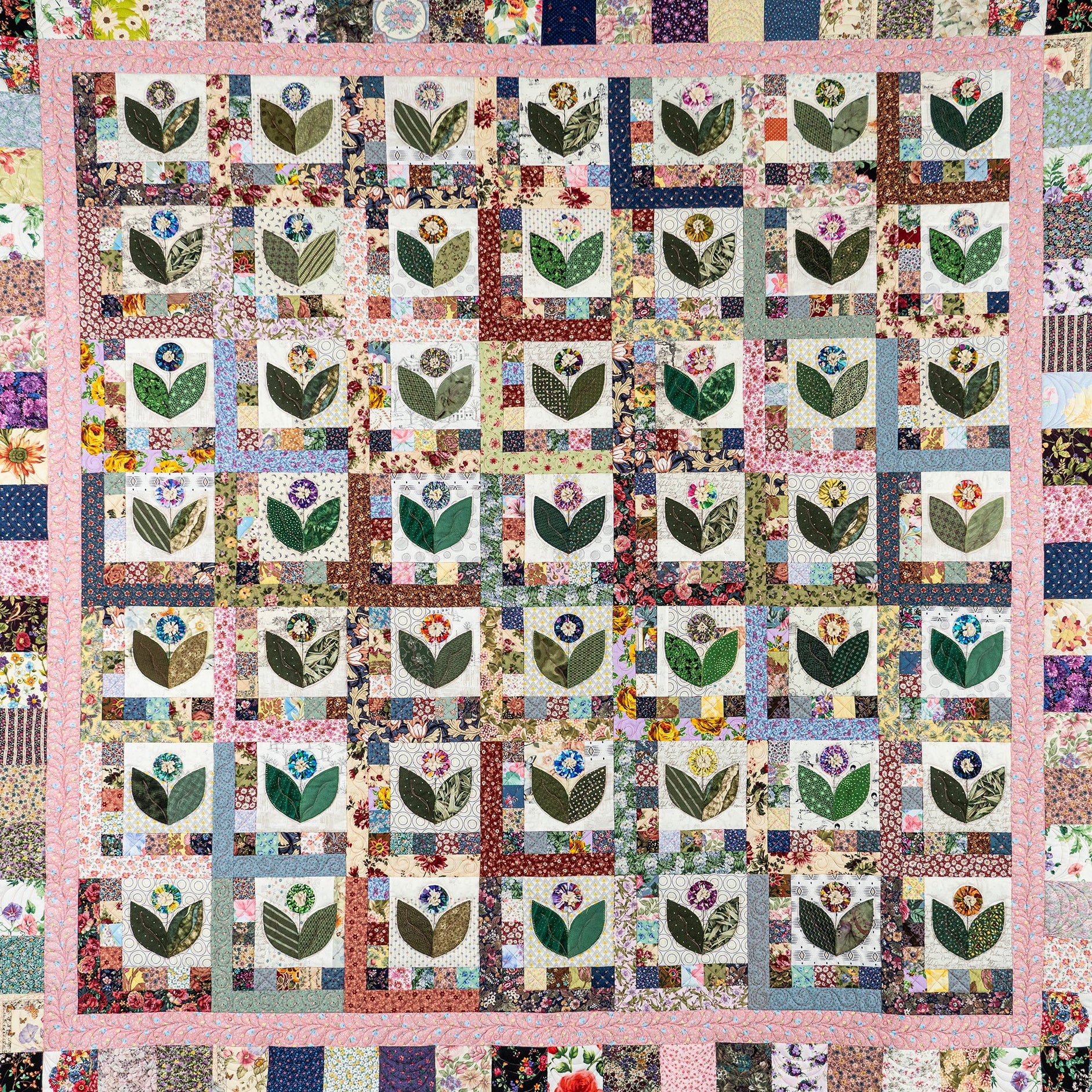 A square patchwork quilt laid flat, featuring a grid of tulip motifs in floral fabric squares, surrounded by a pink border and multicolored patterned edge patches.