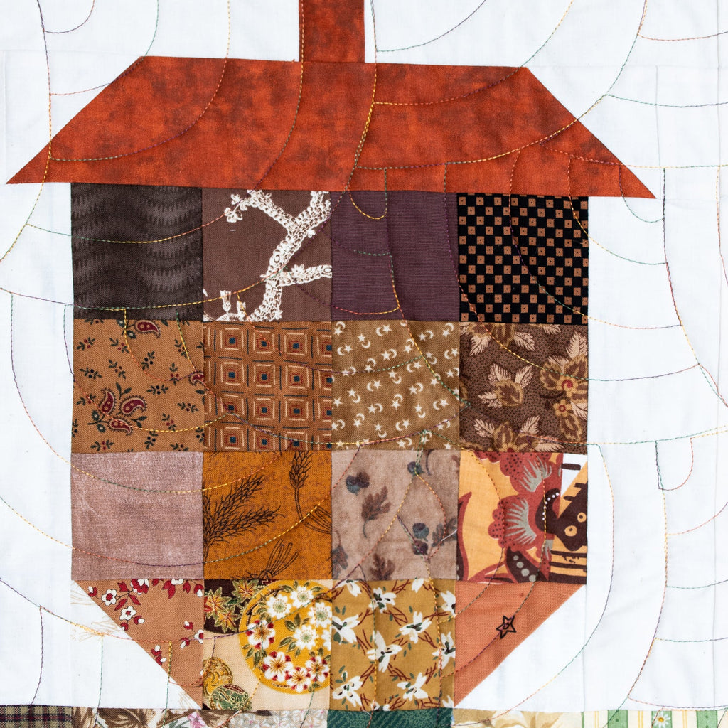 A close-up of a quilted patchwork design shaped like an acorn, made from multiple fabric squares in autumn and neutral tones, with a white quilted background.