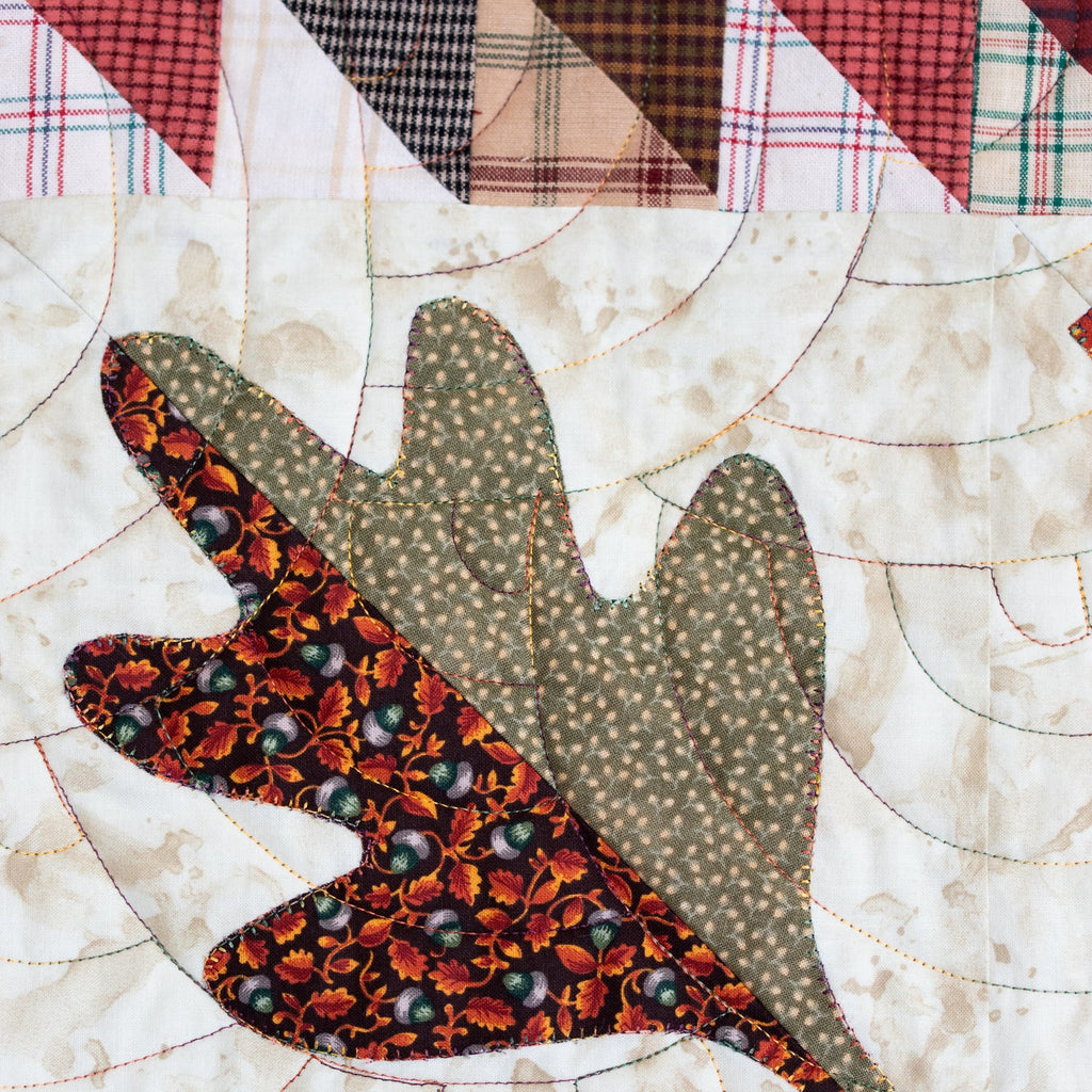 A close-up of a quilted textile featuring a large leaf applique with a red and orange floral pattern on one side and a sage green dotted pattern on the other, set against a cream quilted background with a plaid border.