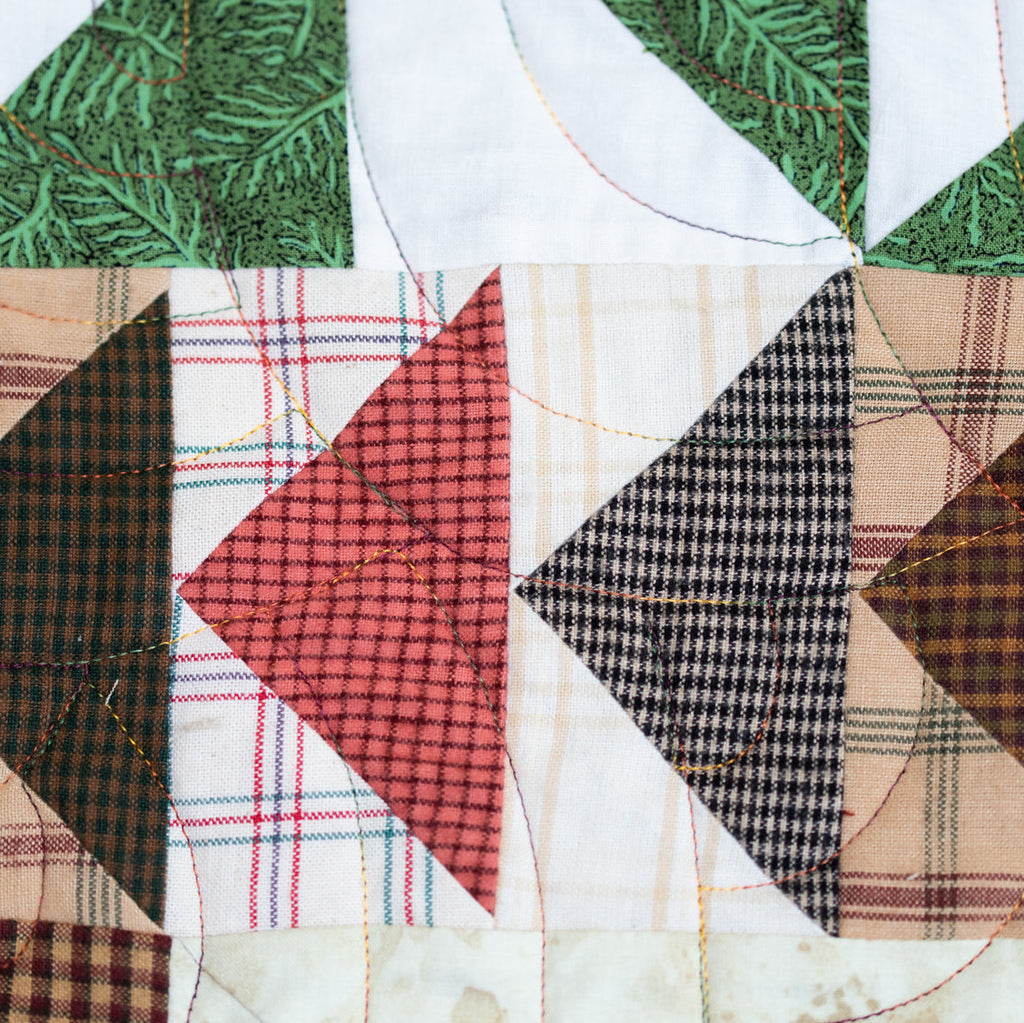 Close-up of a quilted fabric panel with triangular patches in green pine needles, red plaid, black-and-white gingham, and tan plaid on a white background.
