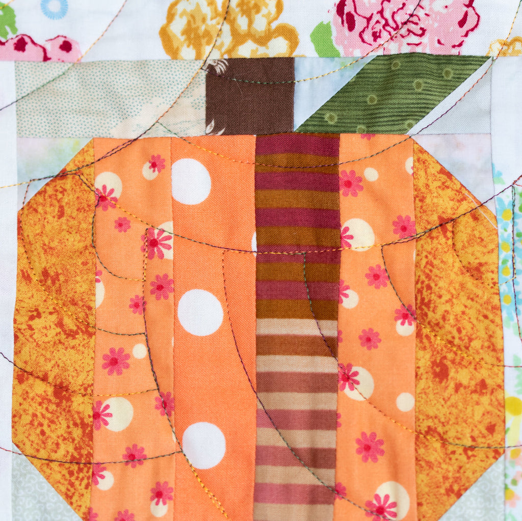 A close-up of a quilted fabric block featuring a stylized pumpkin design. The pumpkin is made of orange, peach, and brown striped and patterned fabric panels with white polka dots and pink floral accents.