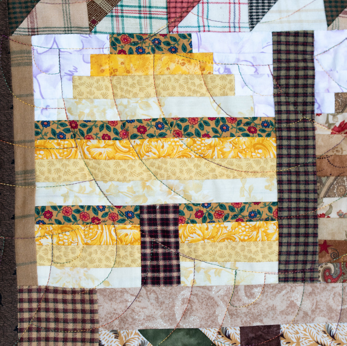 A close-up of a quilted fabric block featuring a stylized tree made of layered yellow, floral, and plaid patterned strips, surrounded by coordinating quilted fabrics.