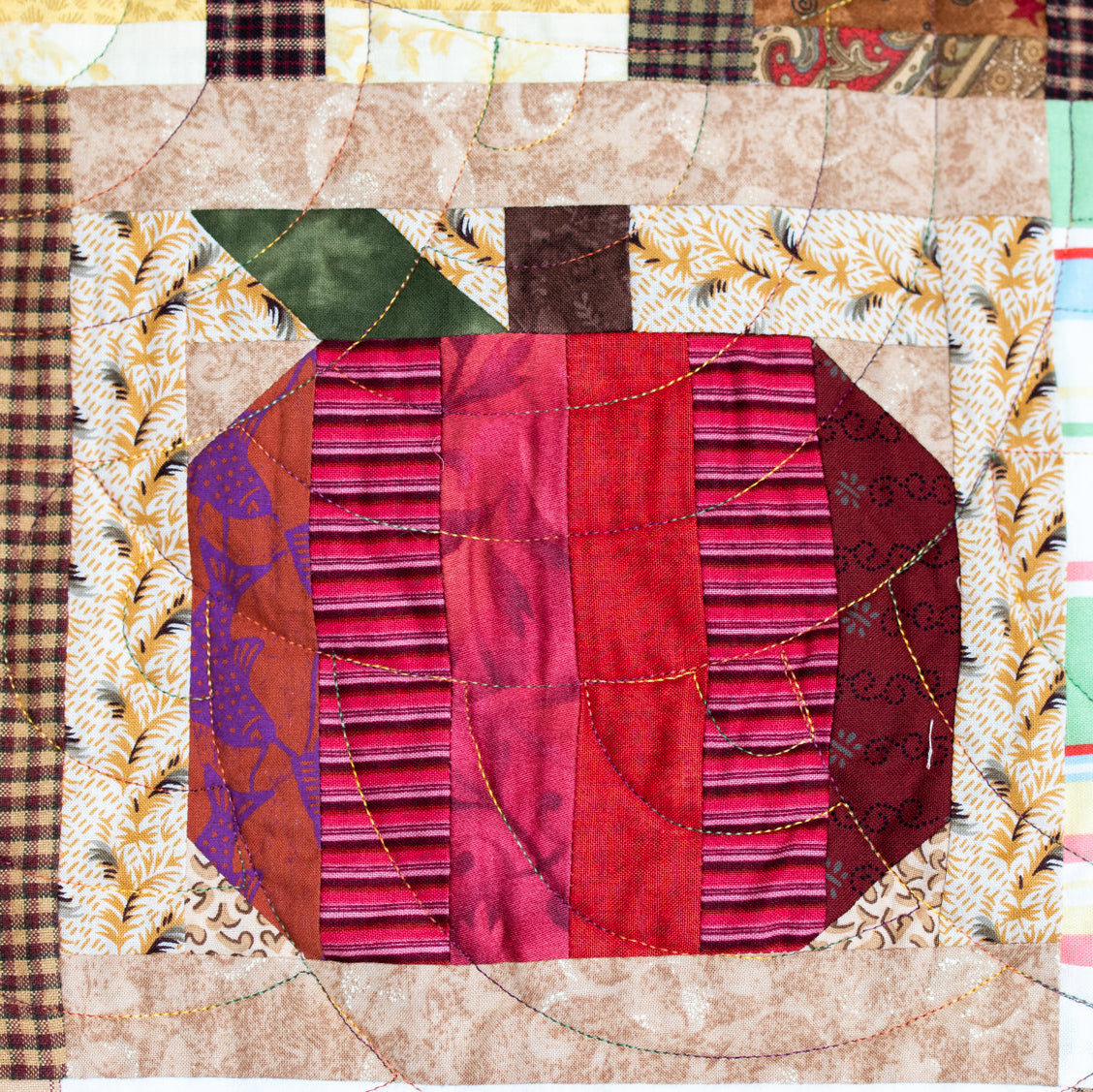 A close-up of a quilted fabric patch featuring a stylized pumpkin design made from red, pink, purple, and brown patterned fabrics, surrounded by cream and brown plaid borders.