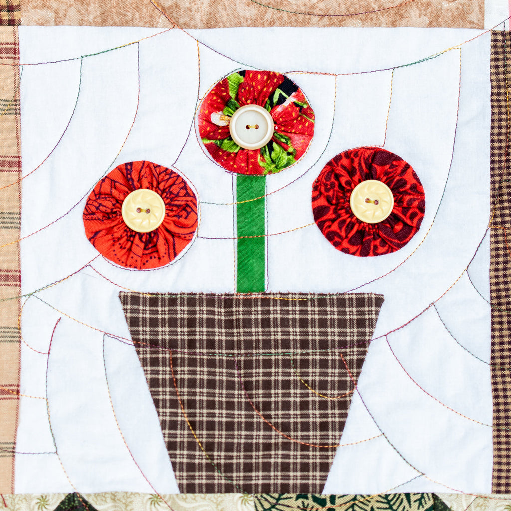A close-up of a handmade quilted fabric art piece depicting three stylized red flowers in a brown plaid pot on a white quilted background.