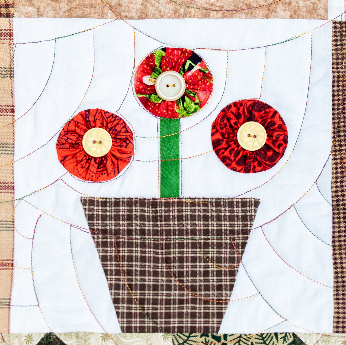 A close-up of a handmade quilted fabric art piece depicting three stylized red flowers in a brown plaid pot on a white quilted background.