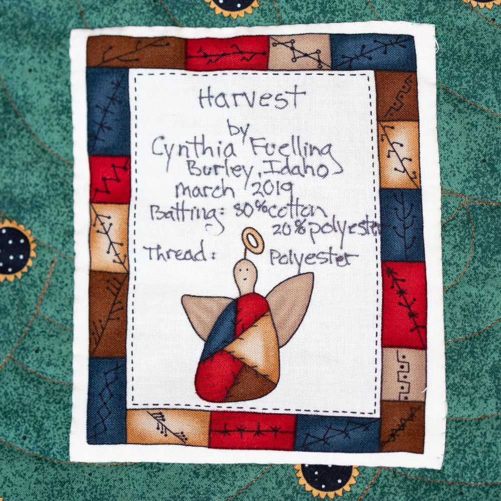 A handmade quilted fabric patch with a stitched white center panel reading 'Harvest by Cynthia Fuscella' with a hand-drawn angel figure, bordered by a patchwork design in red, blue, brown, and cream, laid flat on a green quilted textile background.