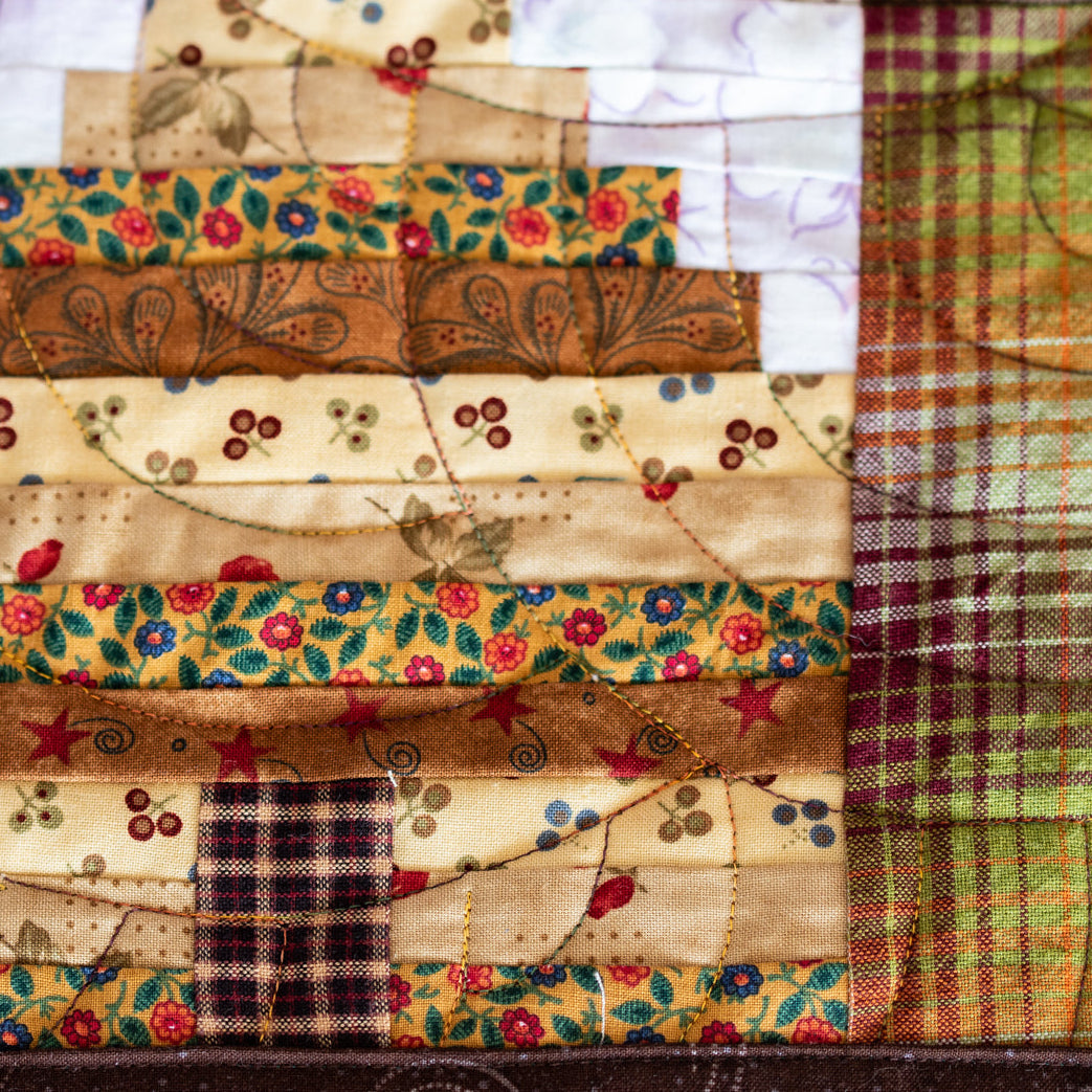 A close-up of a quilted fabric patchwork design, featuring a tree silhouette made of layered patterned fabric strips in beige, brown, and floral prints, next to a green and orange plaid panel.