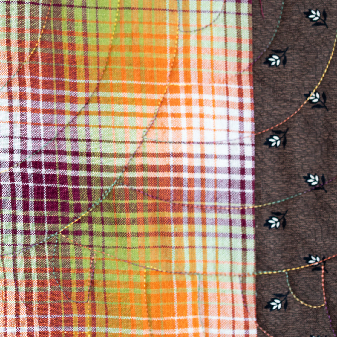 Close-up of a quilted fabric with a plaid pattern in orange, yellow, green, and purple on the left, and a solid dark brown section on the right with embroidered black and white floral details.