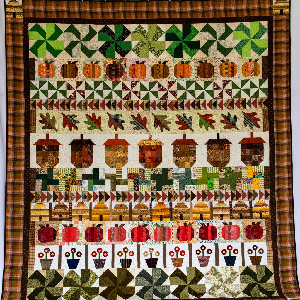 A square patchwork quilt hanging flat against a white backdrop. The quilt features autumn-themed rows with green clovers, orange and brown pumpkins, red acorns, fall leaves, acorns, and acanthus leaves, all framed by a brown and gold plaid border.