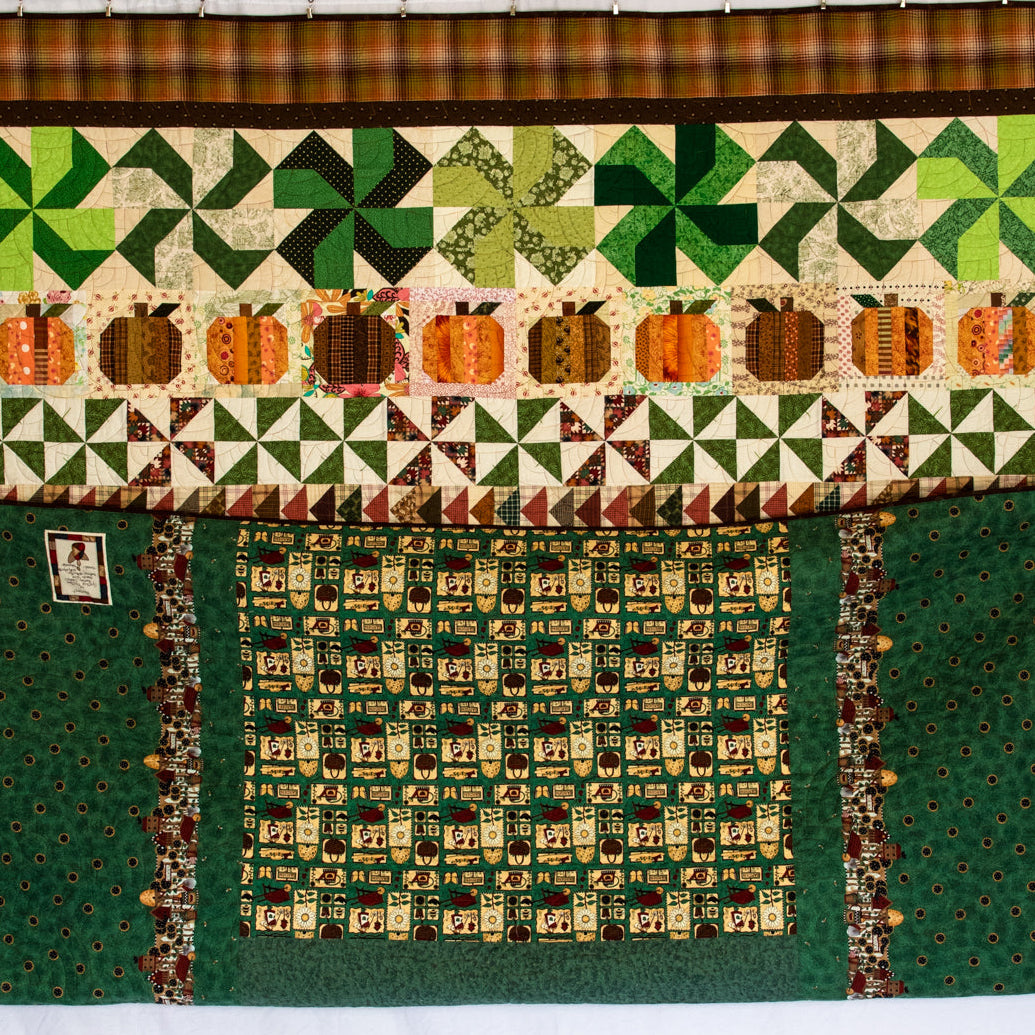 A large quilted textile piece displayed flat against a white backdrop, featuring autumn-themed patchwork sections including green and orange pumpkins, pinewheel patterns, floral clovers, and dark green borders.