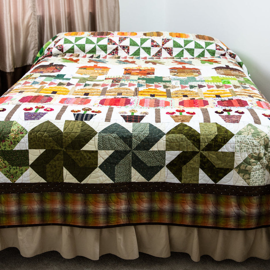 A colorful quilted bedspread with a ruffled skirt, featuring a patchwork design of autumn leaves, pumpkins, pinecones, and geometric patterns in green, orange, red, yellow, and white. The quilt is displayed on a bed in a bedroom setting.