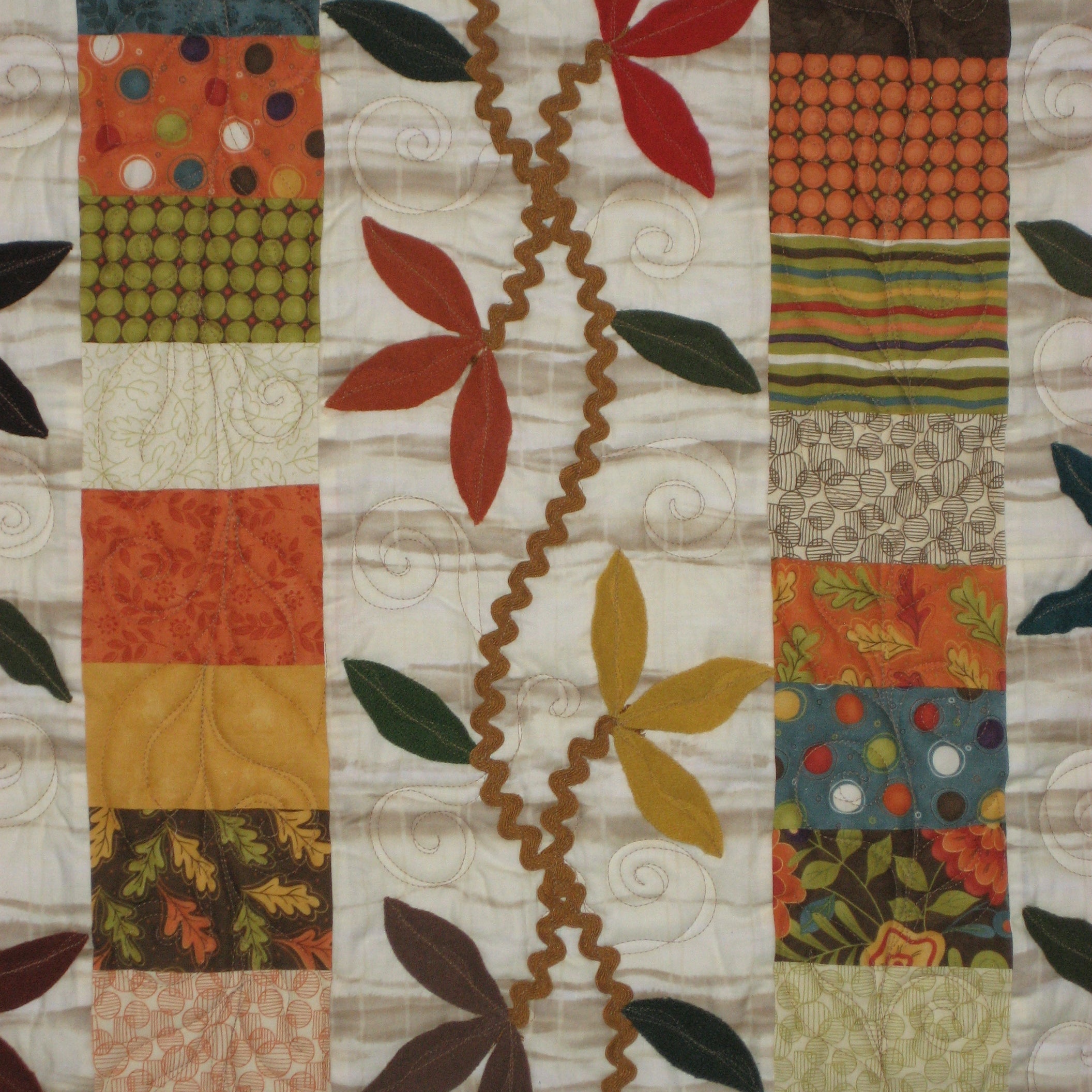 A close-up of a quilted fabric featuring a patchwork border with autumn-themed patterns and a central horizontal design of red, orange, yellow, and green leaves connected by a wavy brown vine on an off-white background.