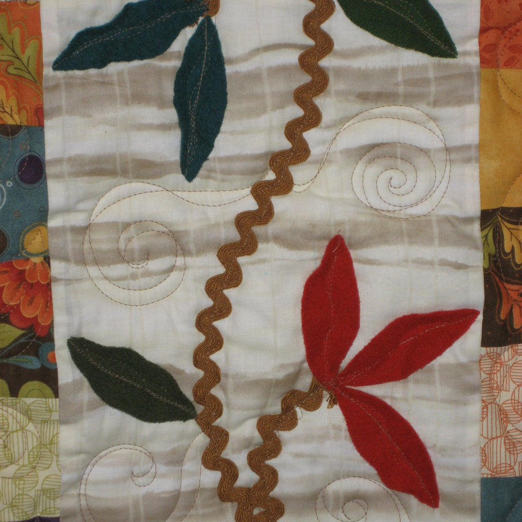 A close-up of a quilted textile featuring a cream background with embroidered swirls, a brown wavy border, and fabric appliques in red, green, and teal. Patchwork borders are visible at the top and bottom.