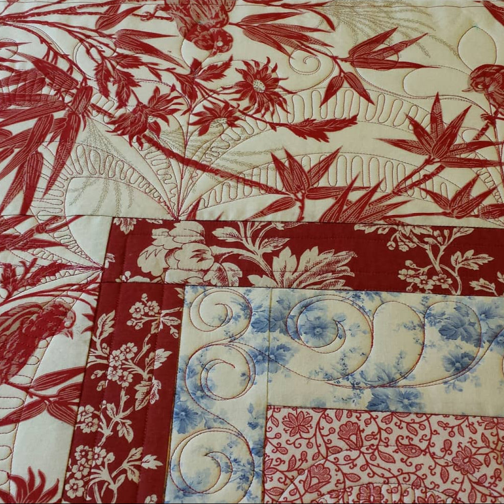 A close-up of a quilted fabric featuring a patchwork design with red and cream botanical prints, including bamboo leaves, flowers, and birds, alongside a blue and cream scroll pattern.