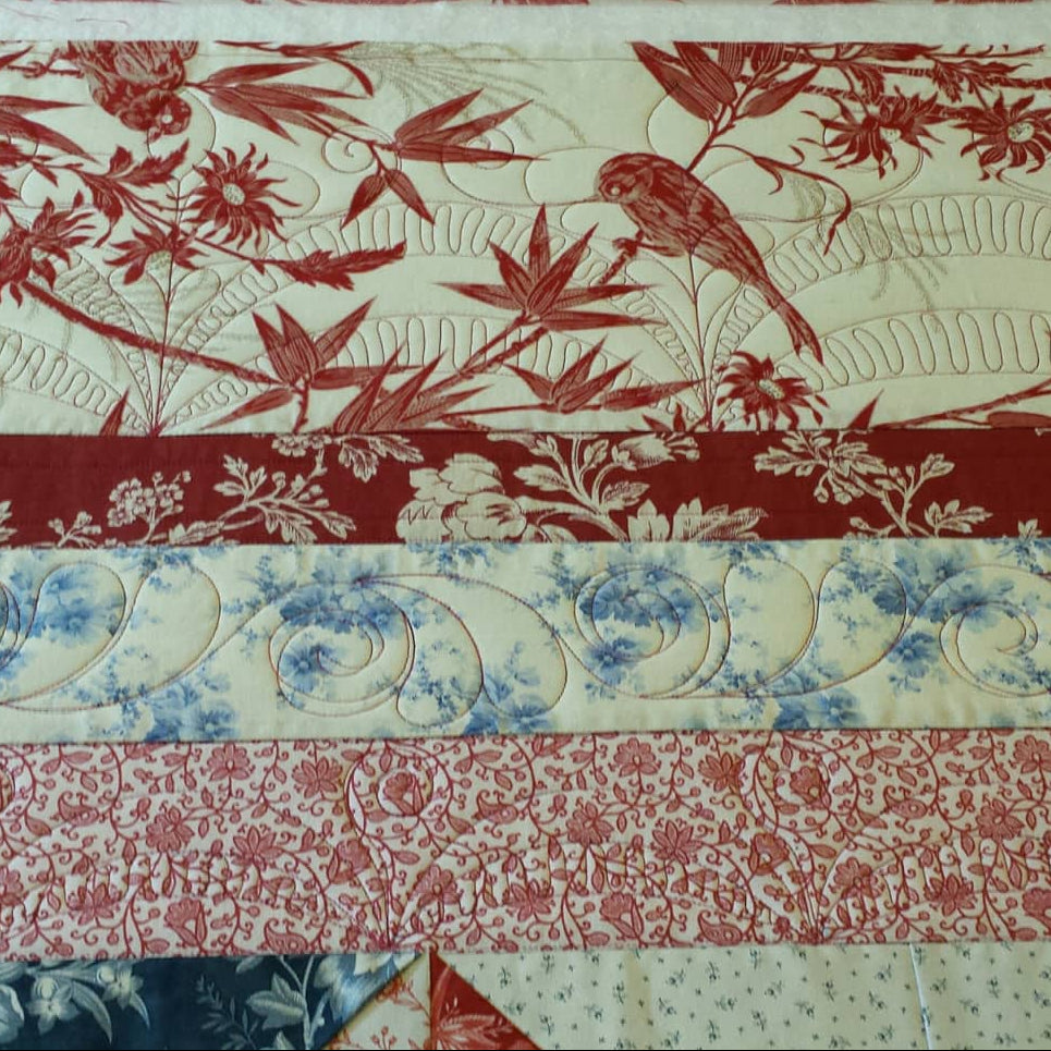 A close-up of a quilted fabric panel featuring multiple horizontal bands of red and blue floral, bird, and scroll patterns on an off-white background.