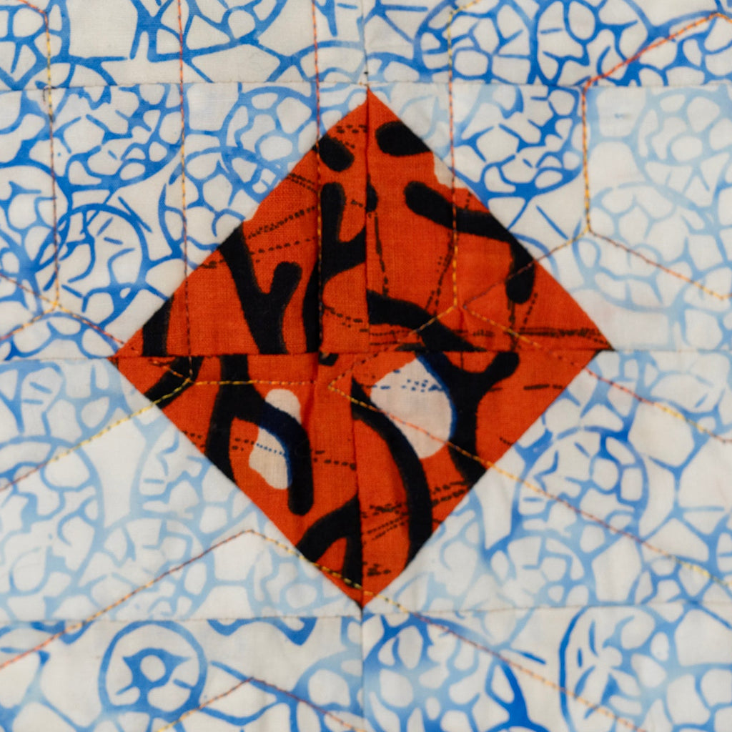 Close up of a quilt block with an orange diamond in the centre with a black print, surrounded by a blue and white abstract print. 