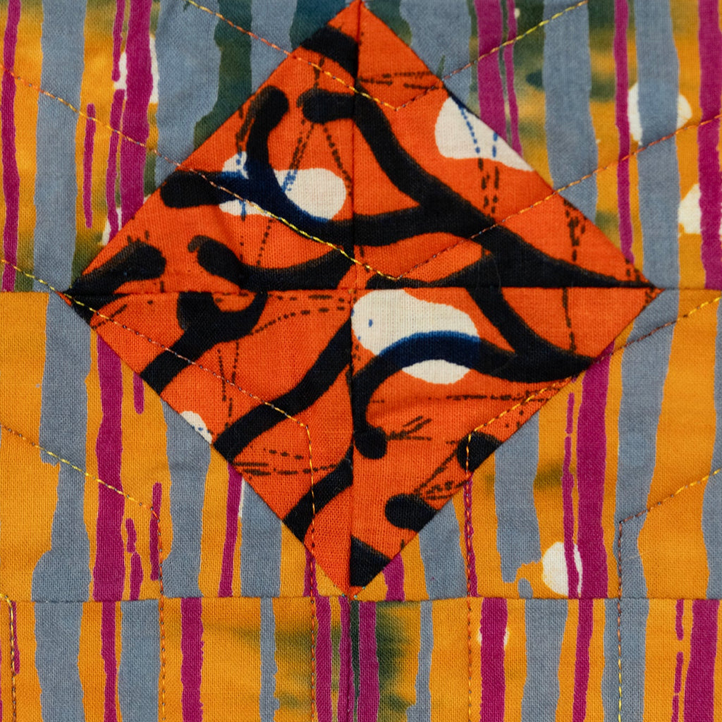 Close up of a quilt block with a orange diamond in the centre with a lack pattern surrounded by a blue, purple and orange striped print.