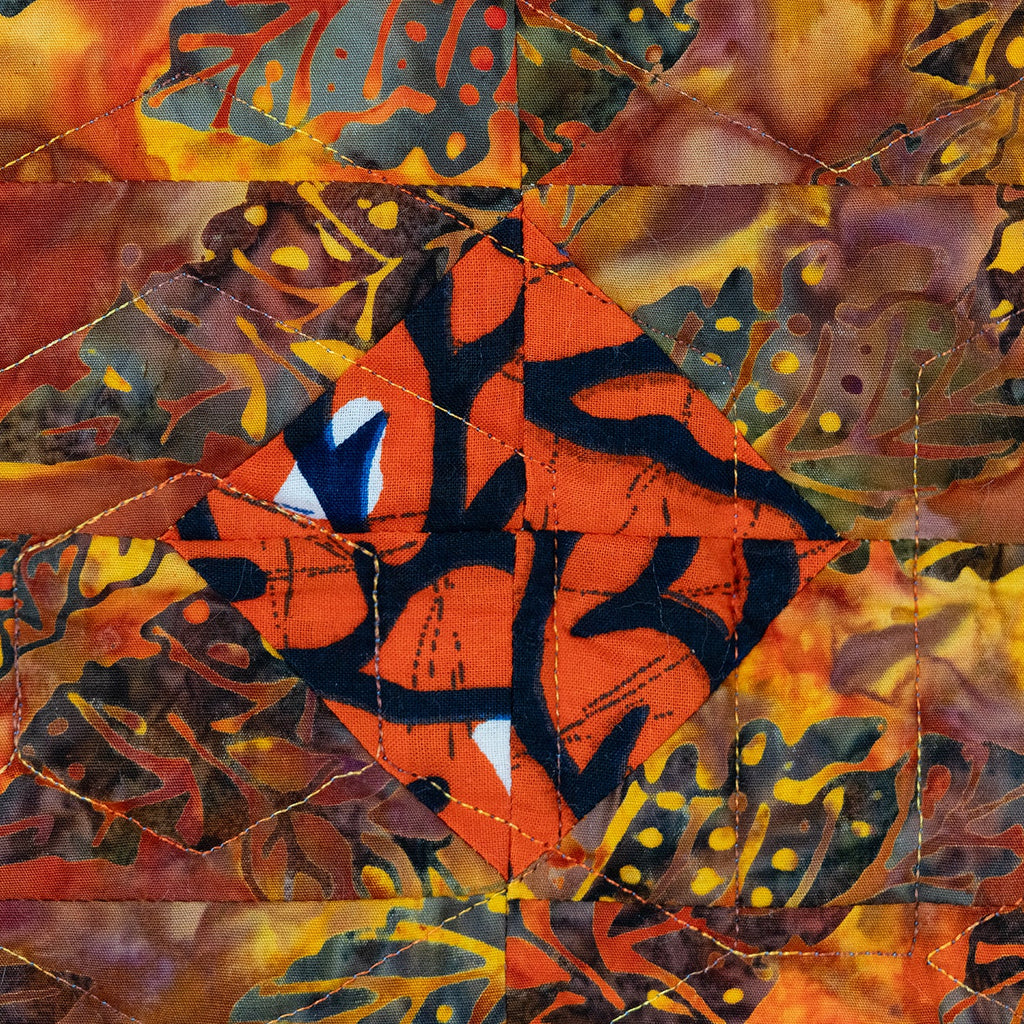 Close up of a quilt block with a orange diamond in the centre with a black print surrounded by a leaf print in autumnal colours. 