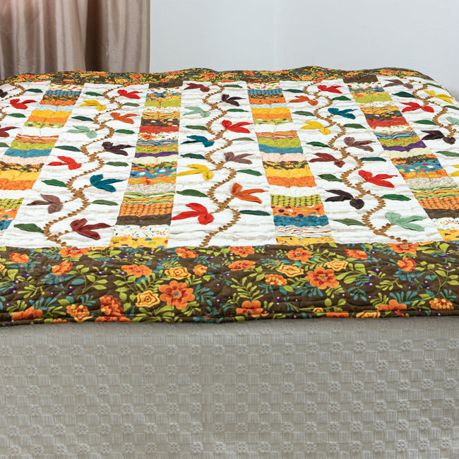 A colorful patchwork quilt with a floral border draped over a bed, shown in a bedroom setting.