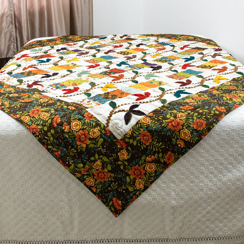 A quilted cream-colored bedspread with fringe trim and a matching floral-patterned throw blanket draped across the top of the bed in a bedroom setting.