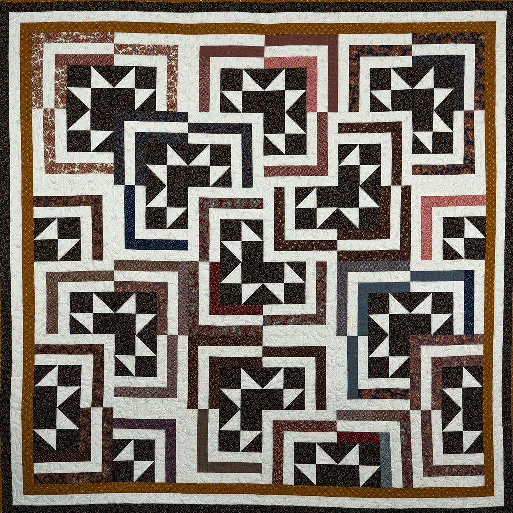 A square quilt with a geometric block pattern in black, white, brown, and muted red, bordered by a wide brown and black frame.