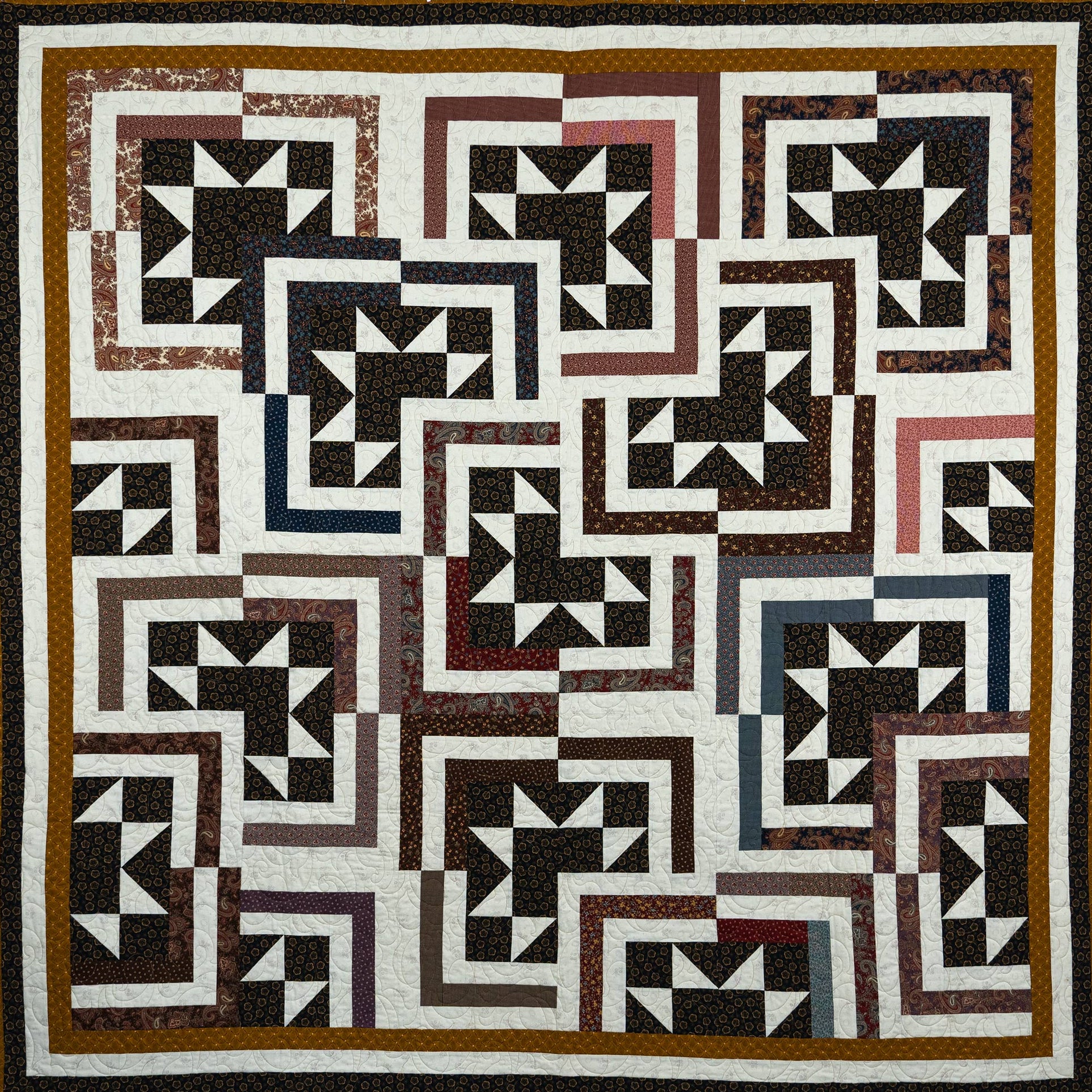 A square quilt with a geometric block pattern in black, white, brown, and muted red, bordered by a wide brown and black frame.