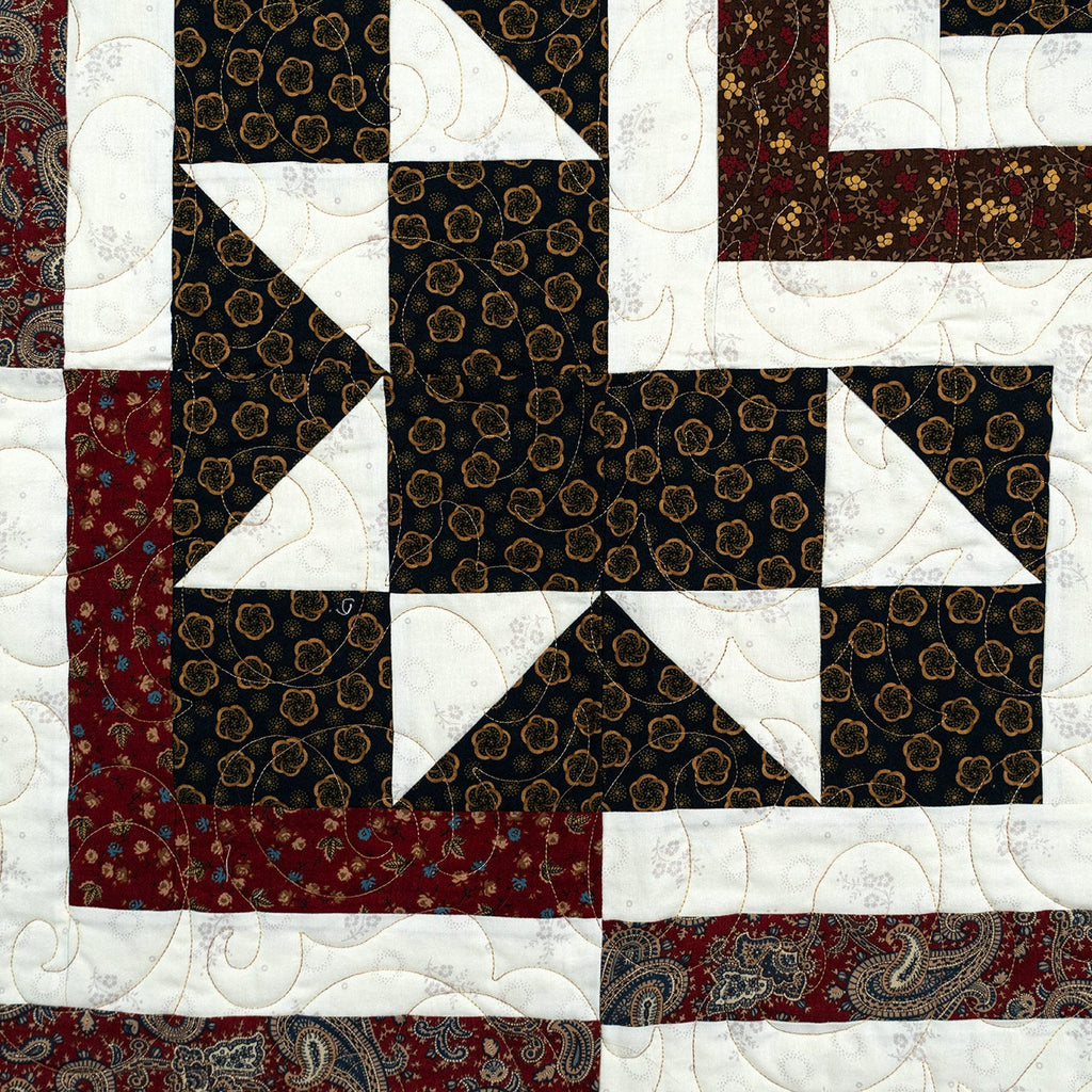 A close-up of a quilted textile with a geometric patchwork design. The pattern includes dark brown and black fabrics with gold floral motifs, solid white sections, and red paisley and floral borders.