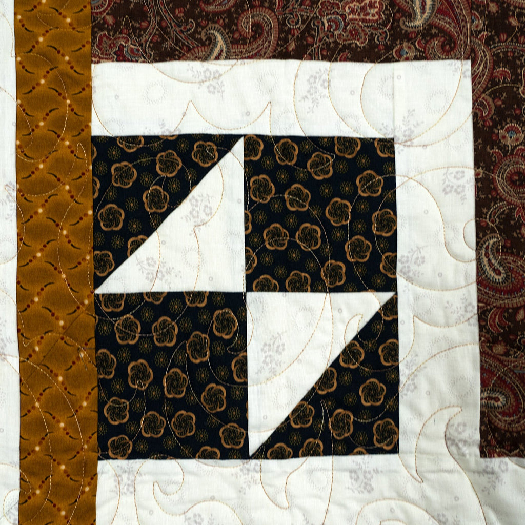 A close-up of a quilted fabric panel featuring a geometric block design with black floral-patterned squares, white triangular sections, a white border, and vertical bands of metallic gold and dark brown paisley fabric.